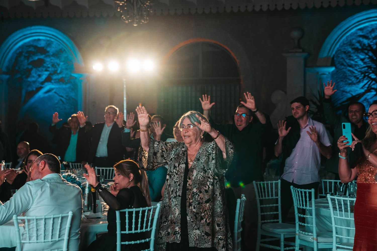 Party guests dance and celebrate with raised arms under blue mood lighting at an elegant venue with white chairs.