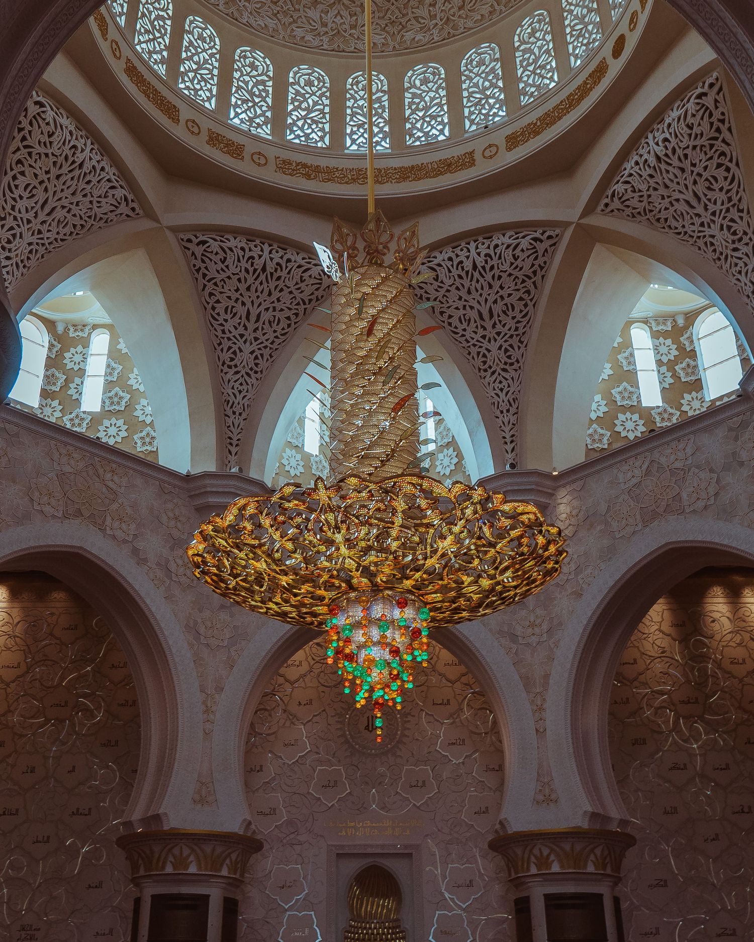 Ornate golden chandelier hangs beneath an intricately decorated dome with Islamic patterns and arched windows in a mosque.