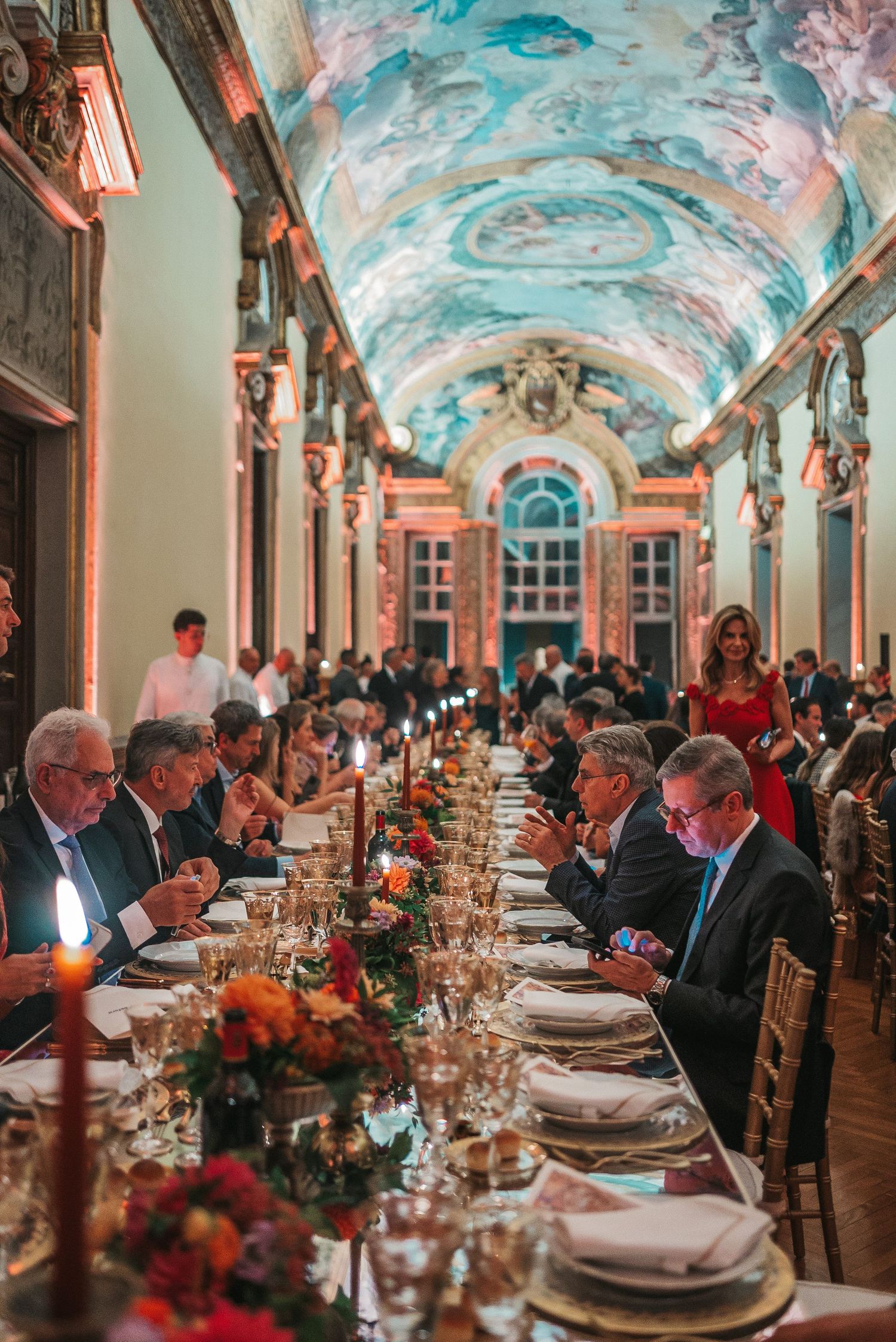 Elegant formal dinner party in ornate hall with vaulted painted ceilings and long tables set with fine china and flowers.