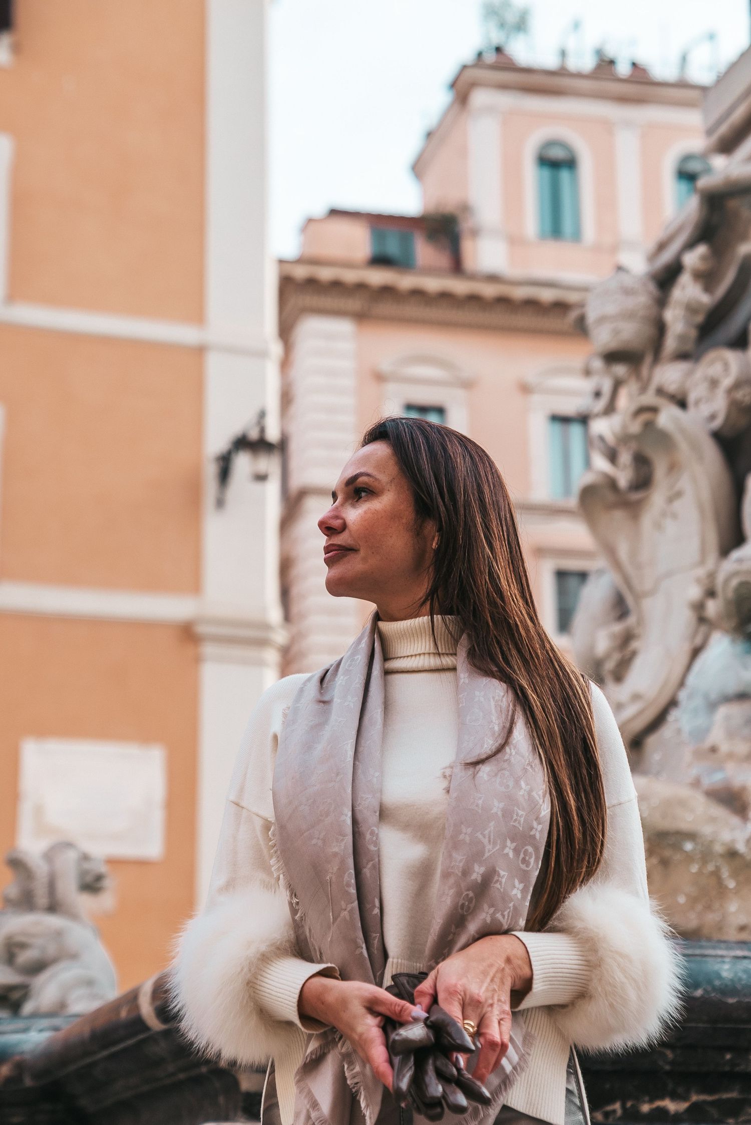 Woman in elegant cream coat with fur trim poses near ornate Roman fountain.