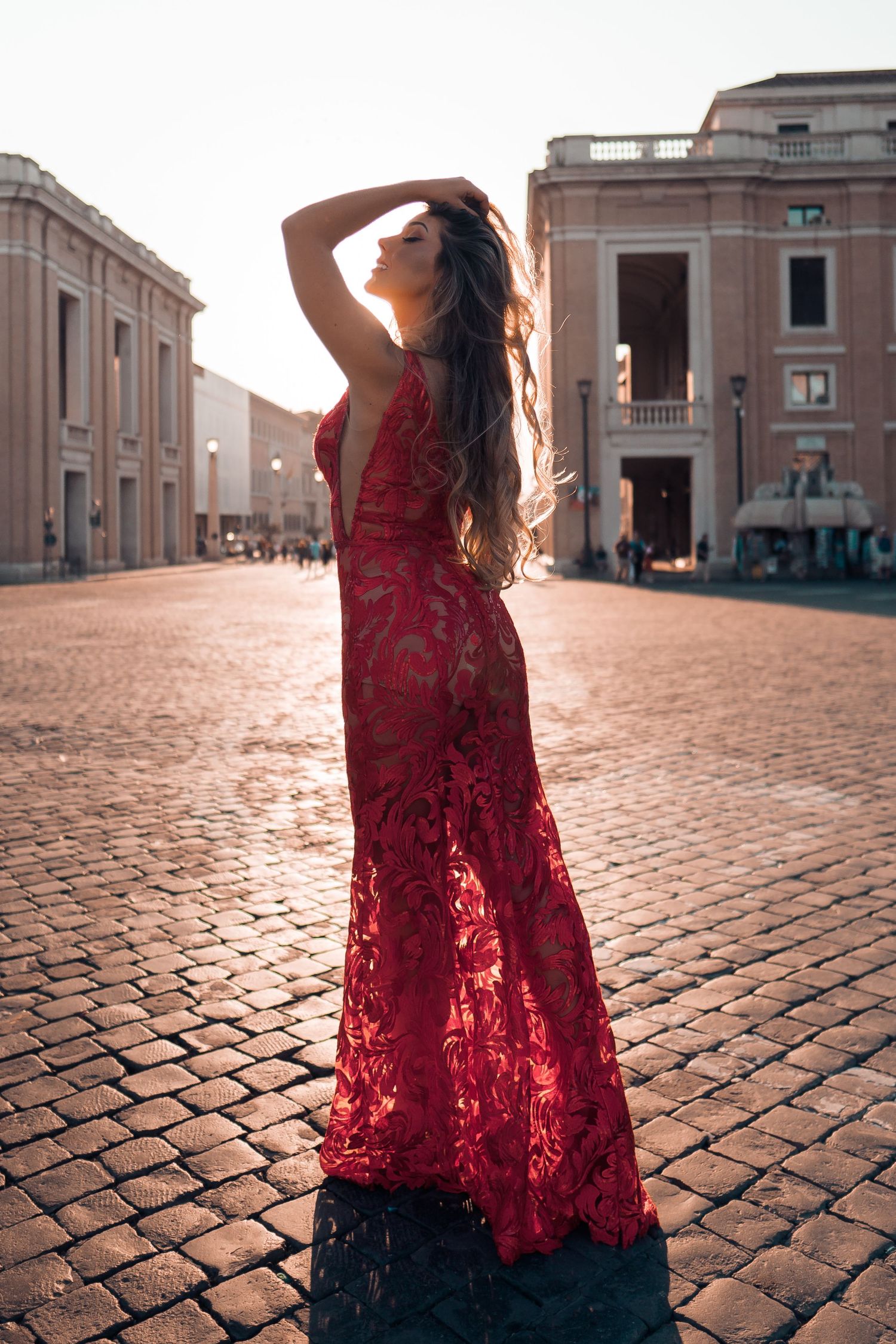 A person in a flowing red lace dress poses on cobblestone streets against a backdrop of classical architecture.