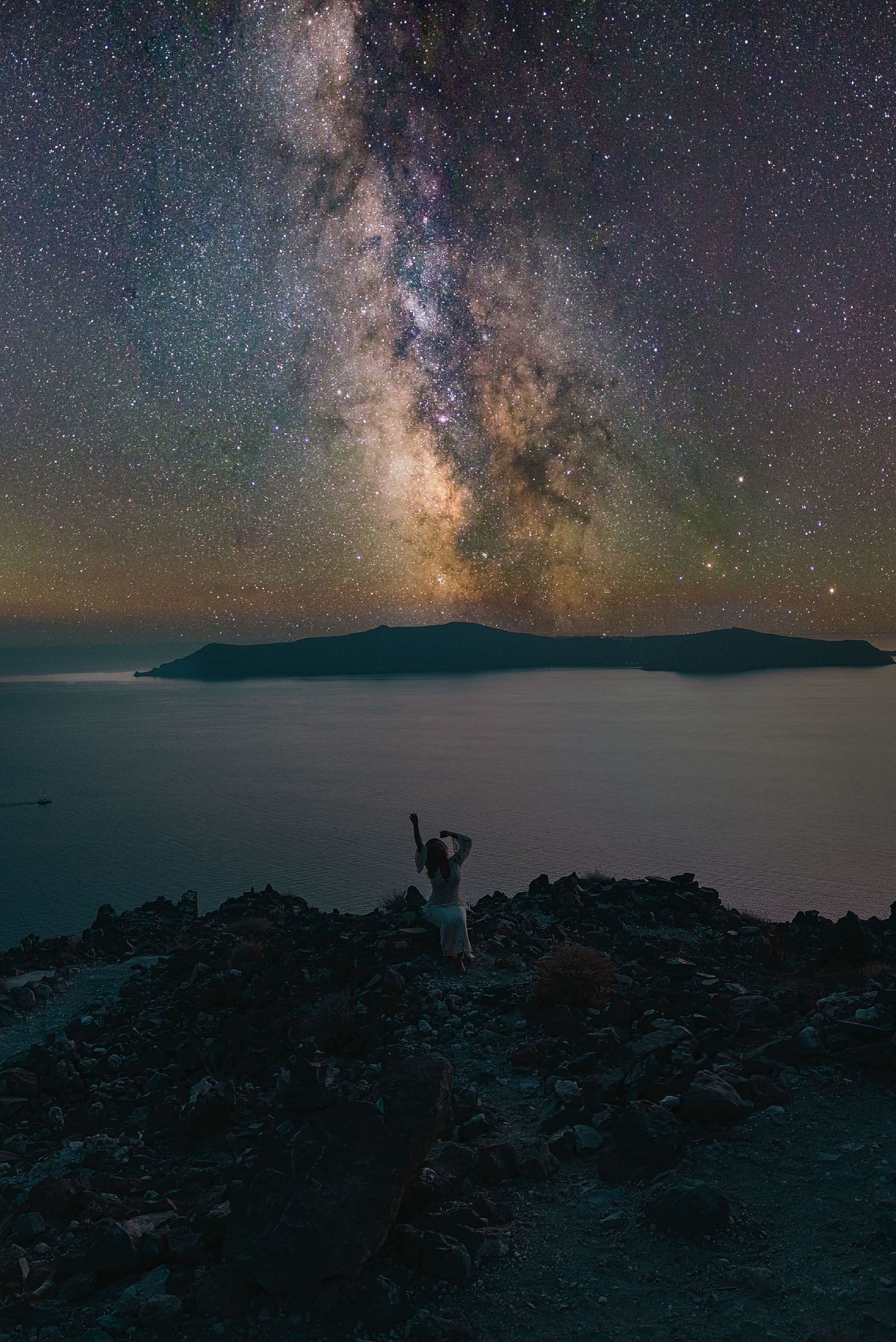 A silhouette stands on a rocky cliff overlooking a lake under the Milky Way galaxy in a stunning night sky.
