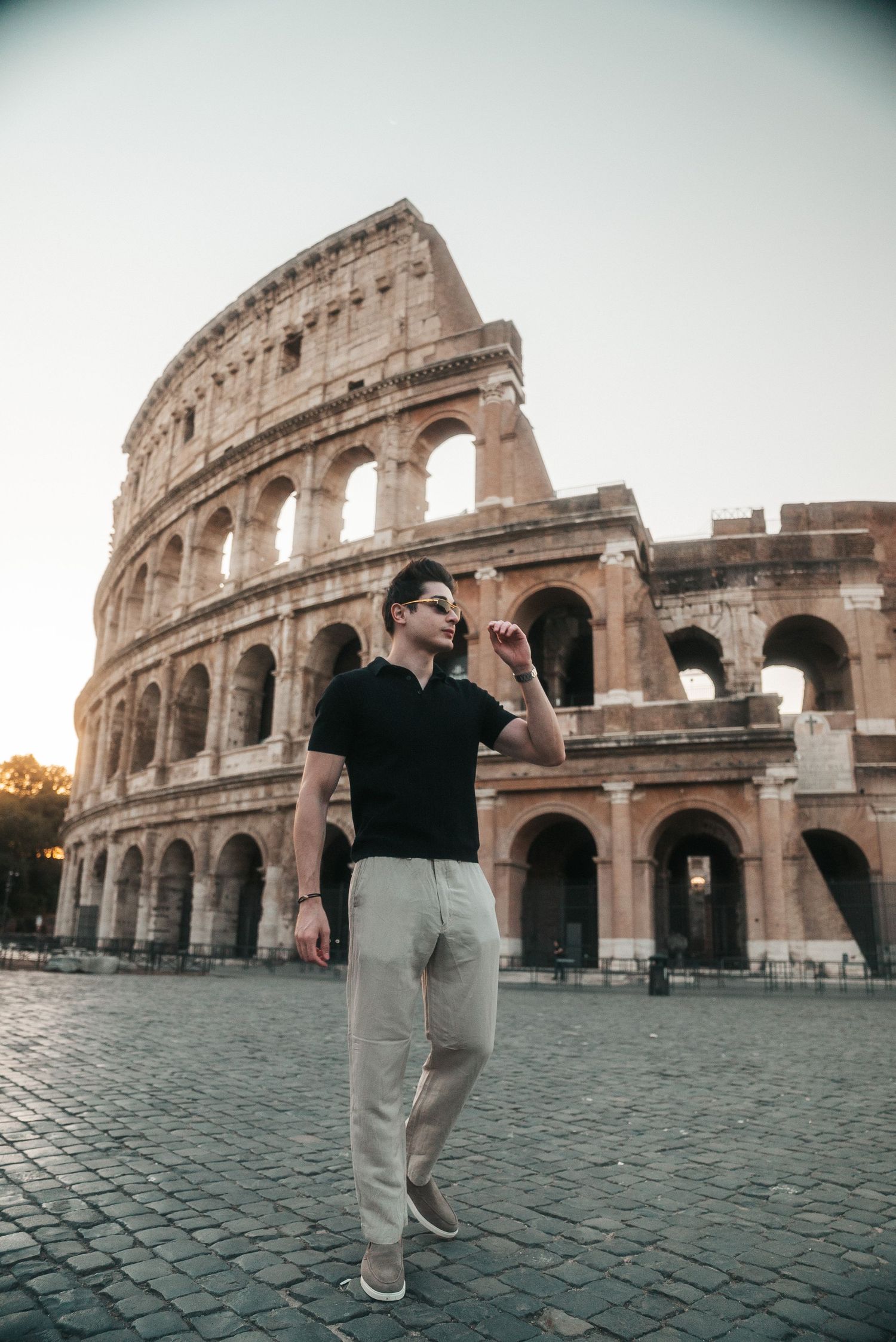 A stylish man in black shirt and beige pants poses confidently in front of the iconic Roman Colosseum at sunset.