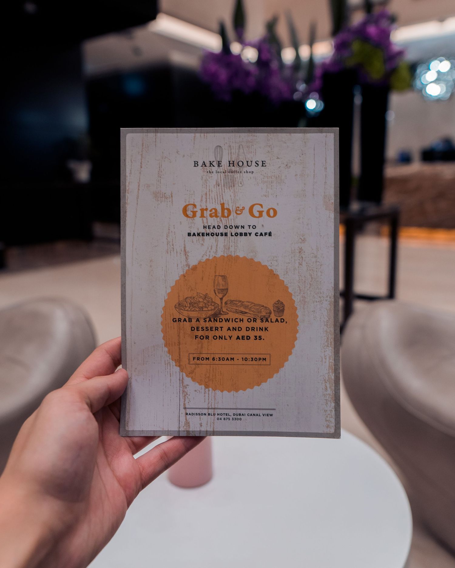A hand holds a Grab & Go menu from Bake House against a blurred background with purple flowers.