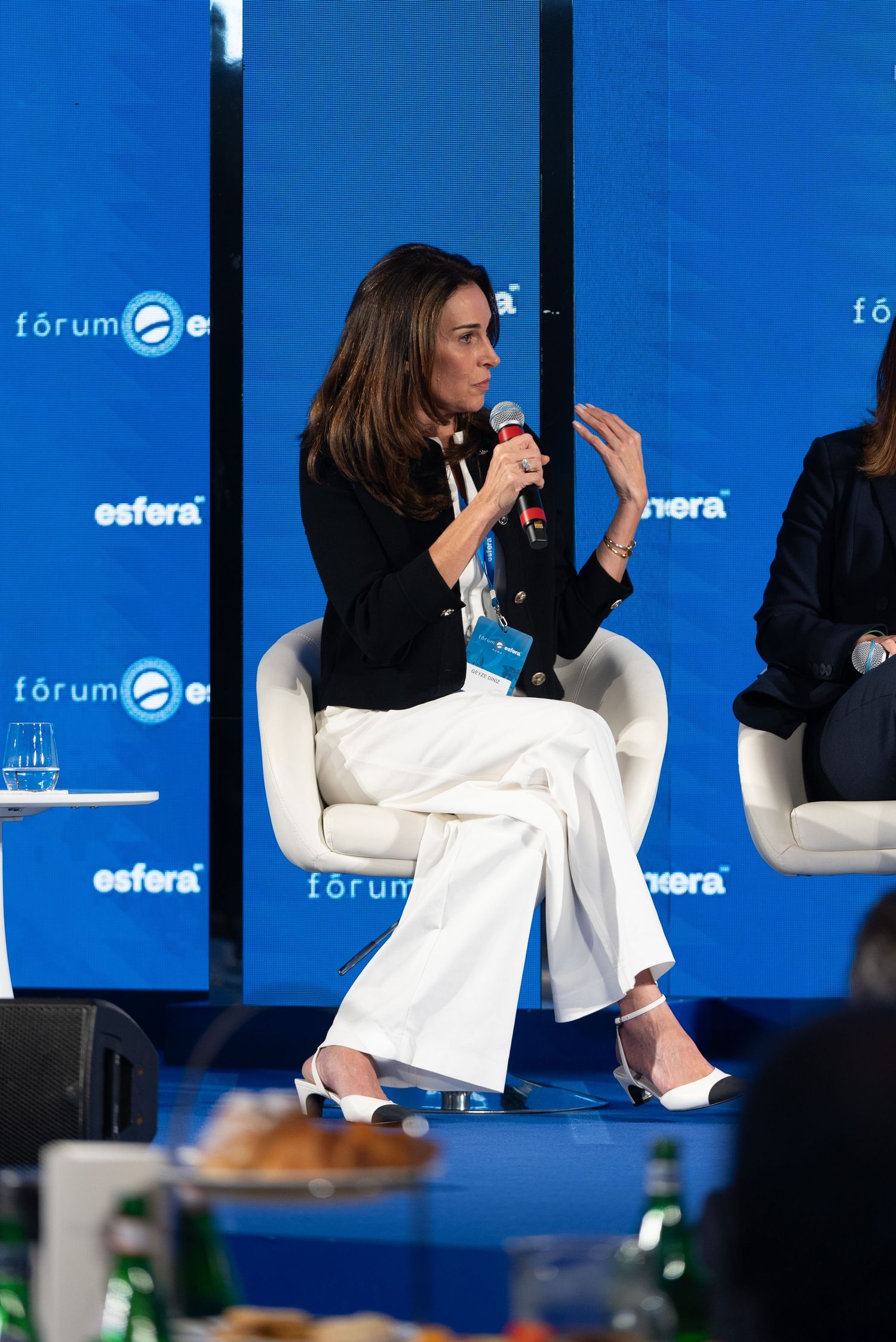 Person in white blazer and dark top gestures while speaking at business forum with blue backdrop and Esfera branding.
