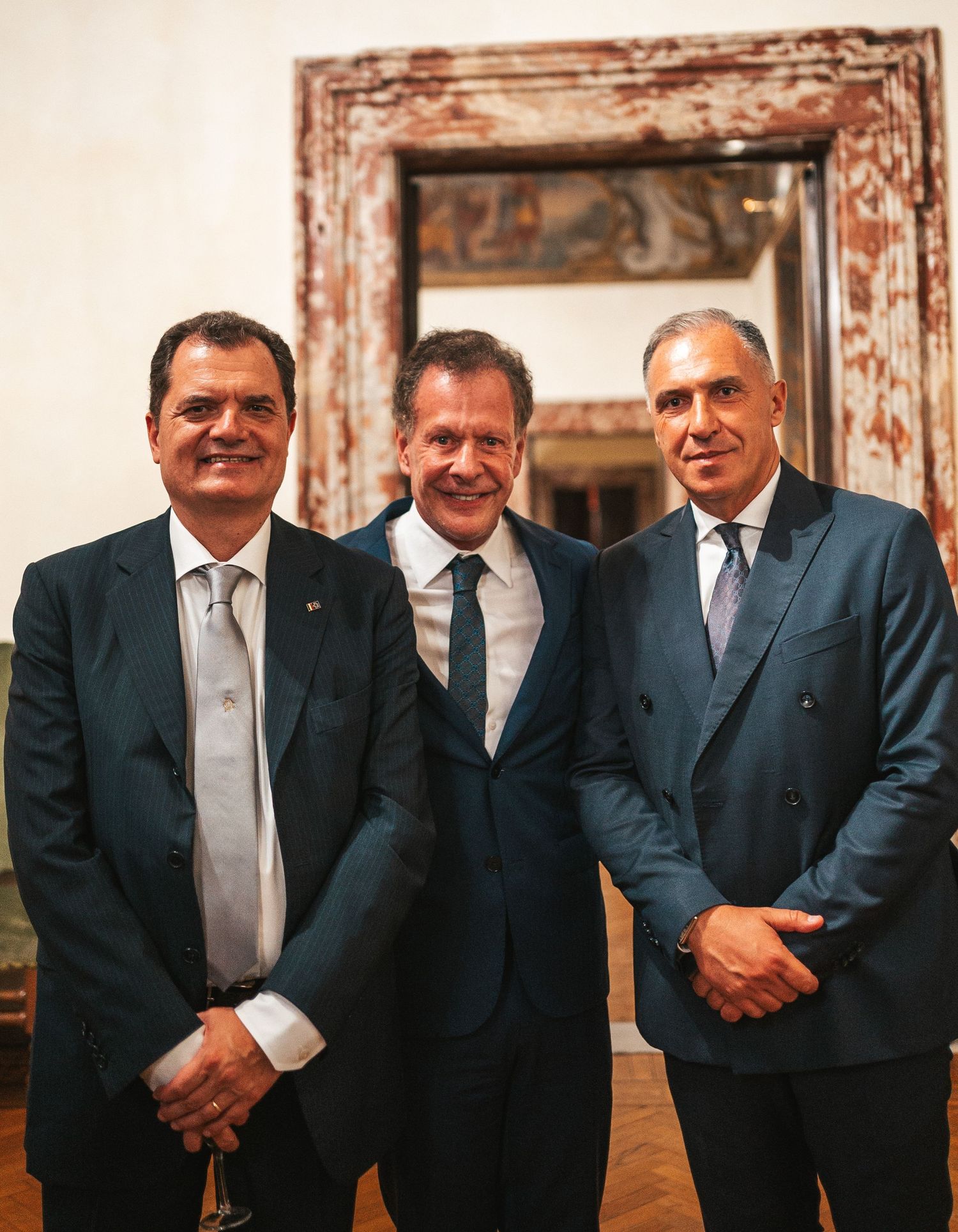 A group of men in dark business suits stand together in a formal setting with ornate wall decorations.