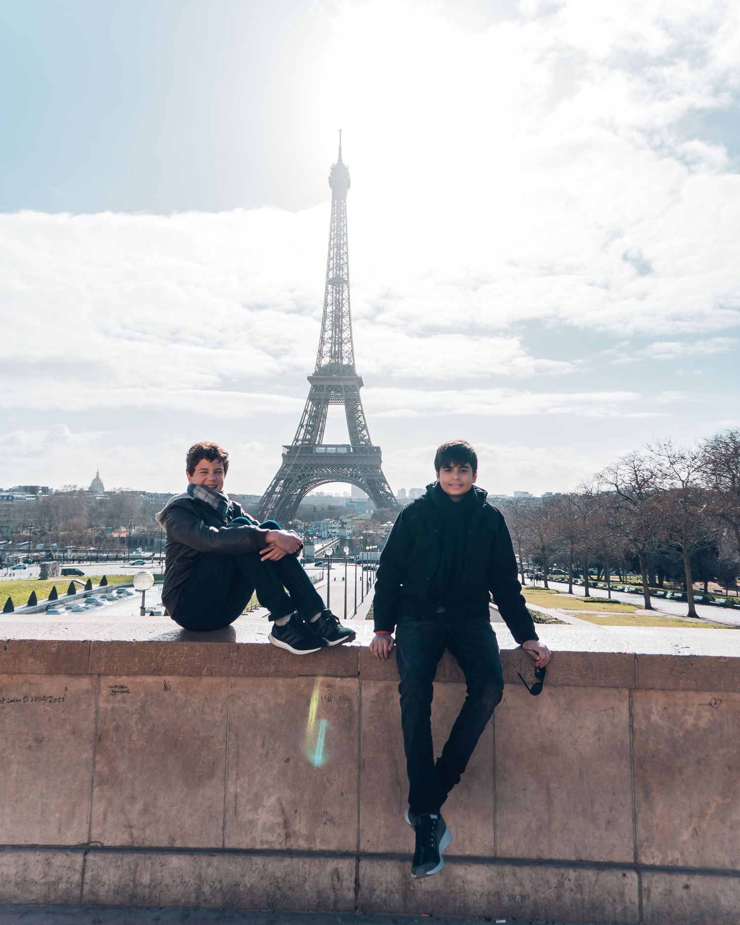 People sitting on a stone wall overlooking the iconic Eiffel Tower in Paris on a cloudy day.