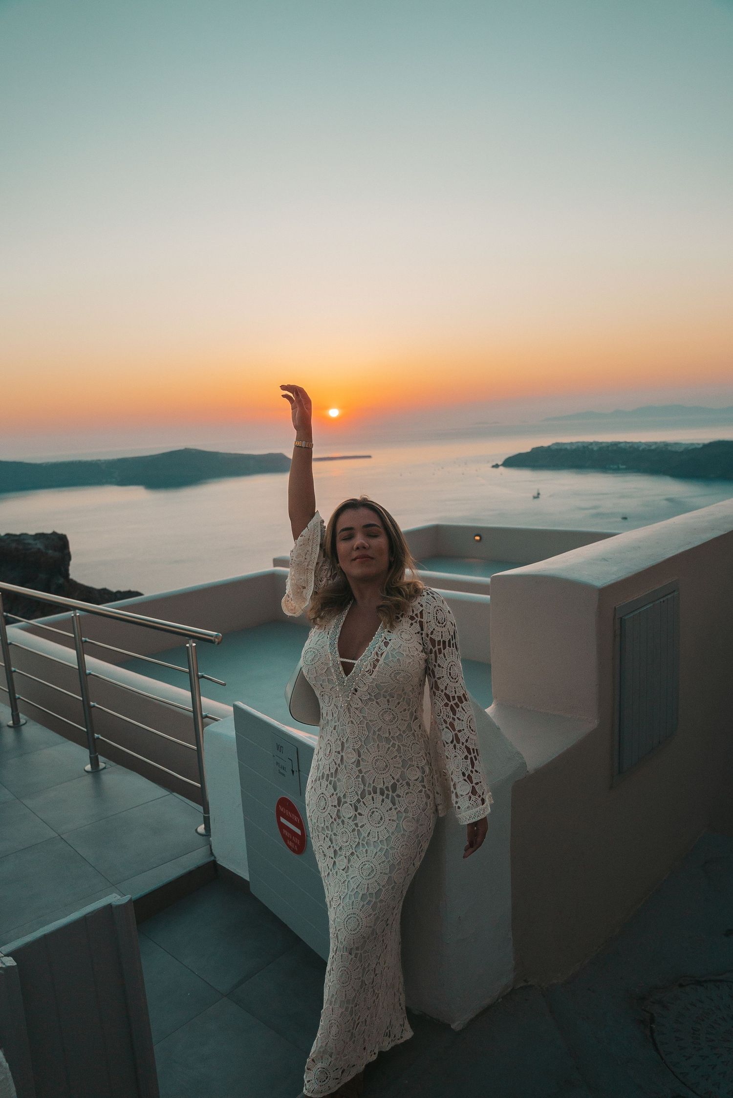 Person in white lace dress poses against stunning Santorini sunset over the Mediterranean Sea and volcanic islands.