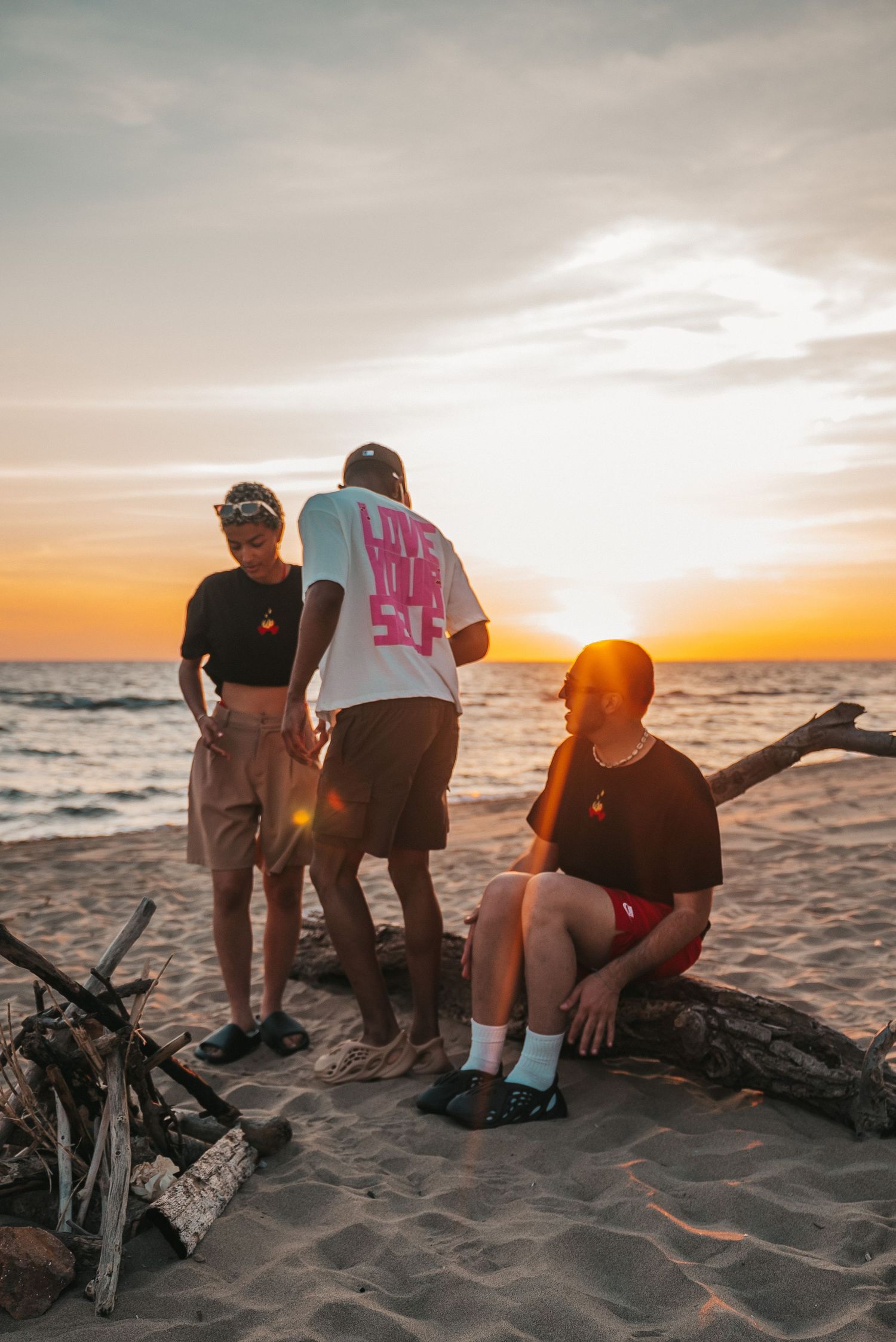 Three friends relaxing on driftwood at the beach during a vibrant orange sunset over the ocean.