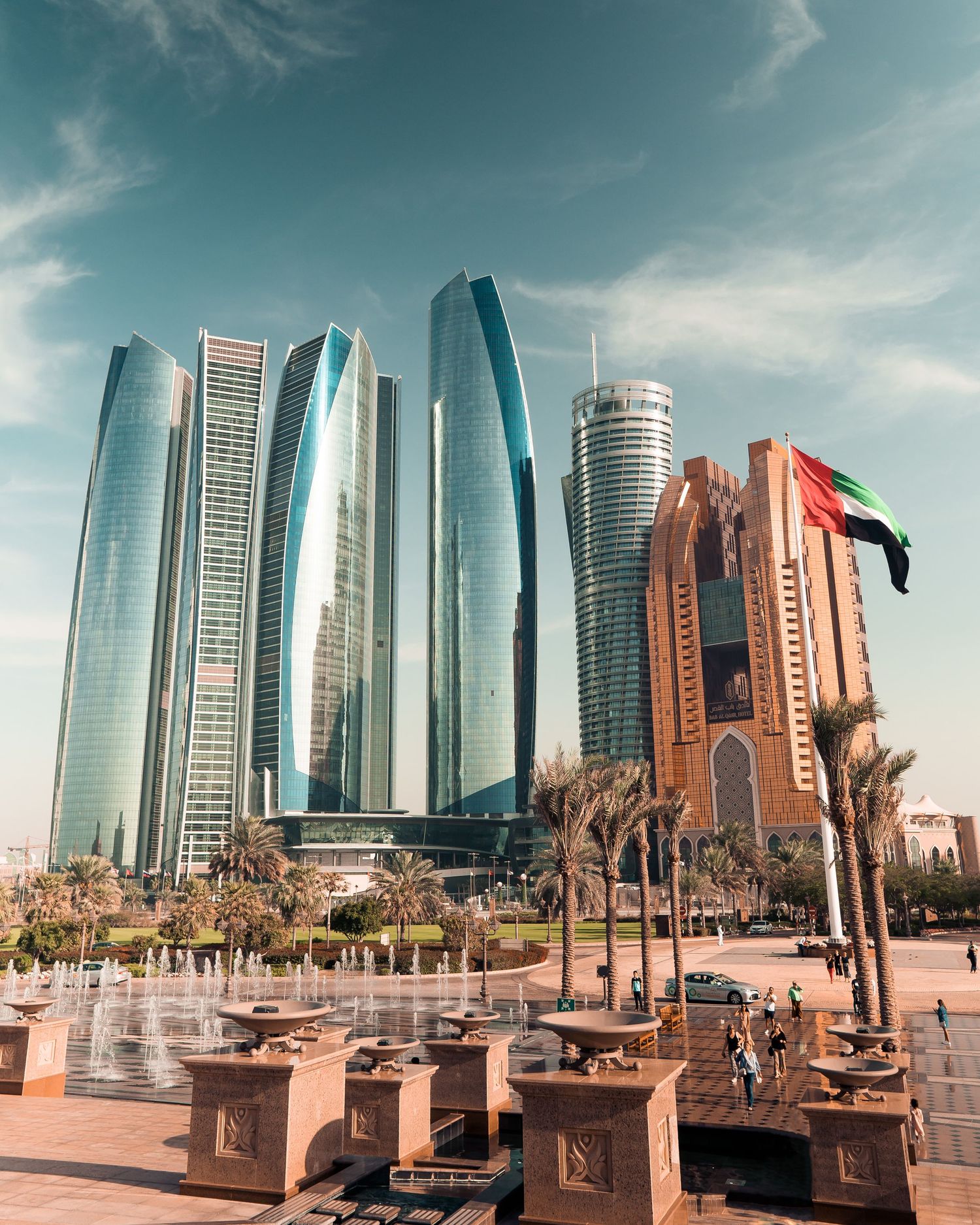 Modern skyscrapers and palm trees line the waterfront promenade in Abu Dhabi's Corniche under a dramatic cloudy sky.