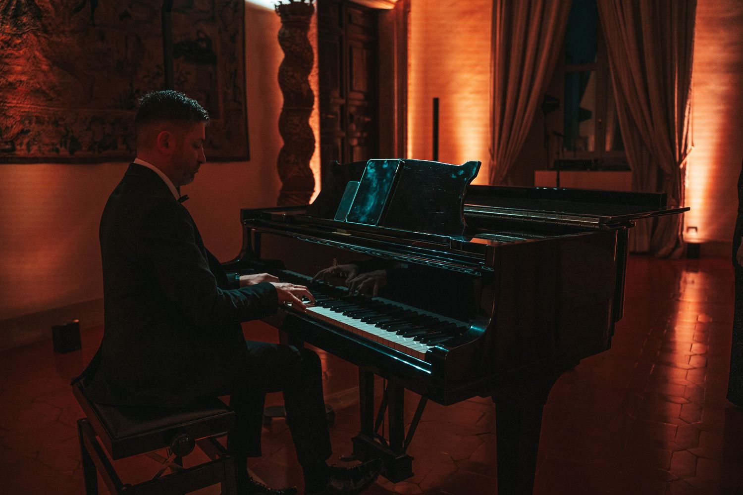 A dark concert hall interior with a grand piano illuminated by warm orange stage lighting and atmospheric shadows.