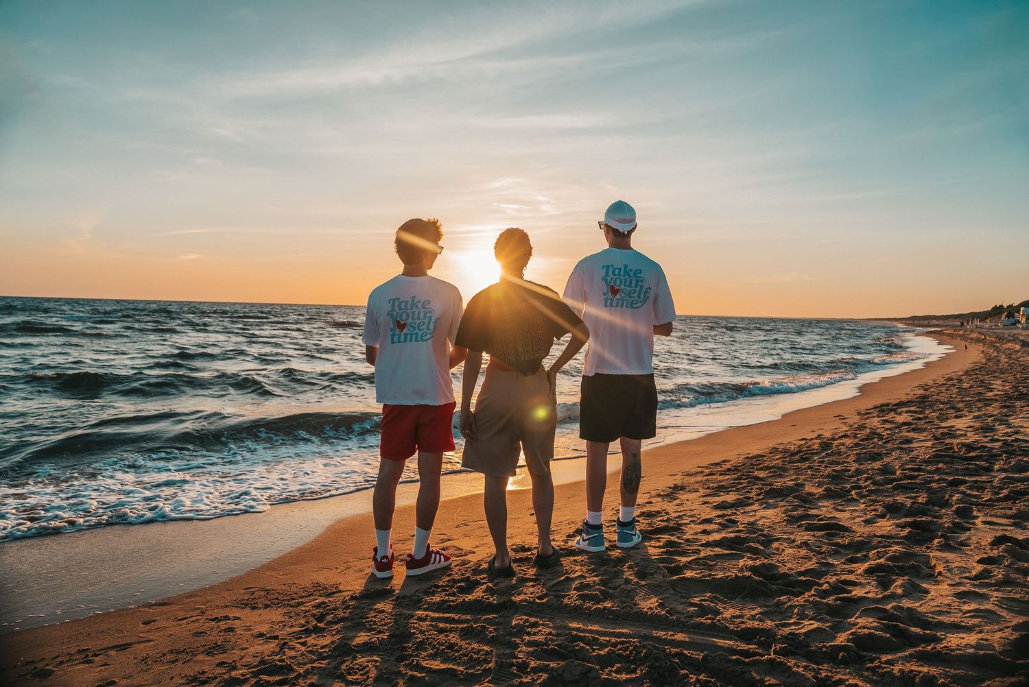 A group of people stand together watching a beautiful sunset over the ocean from a sandy beach.