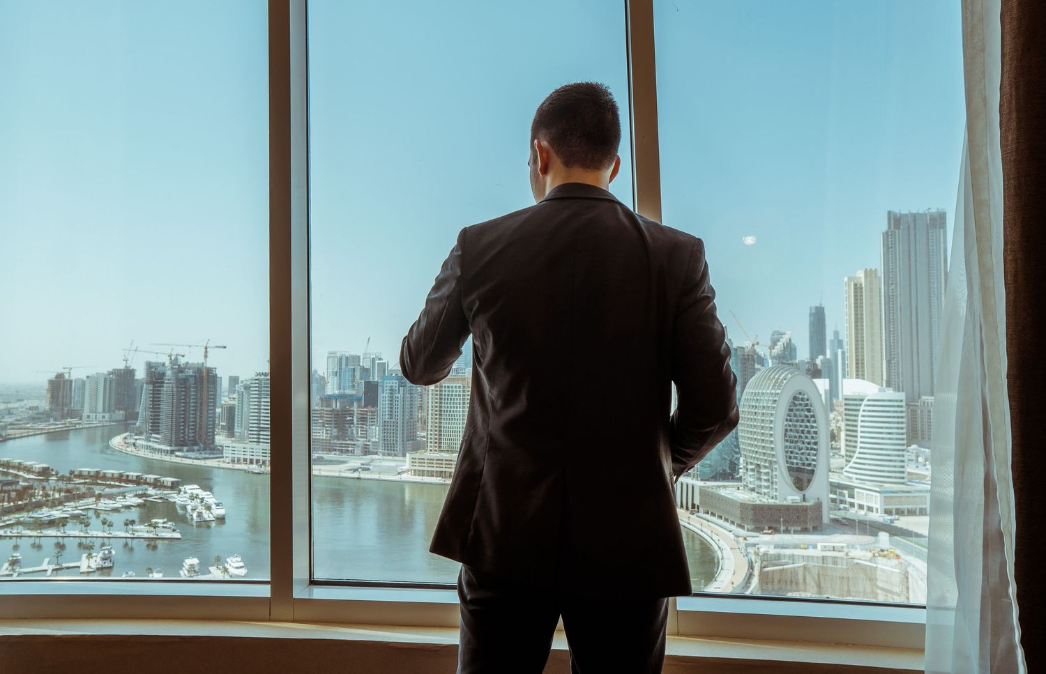 A person stands at a large window overlooking a cityscape with modern skyscrapers and buildings in the background.