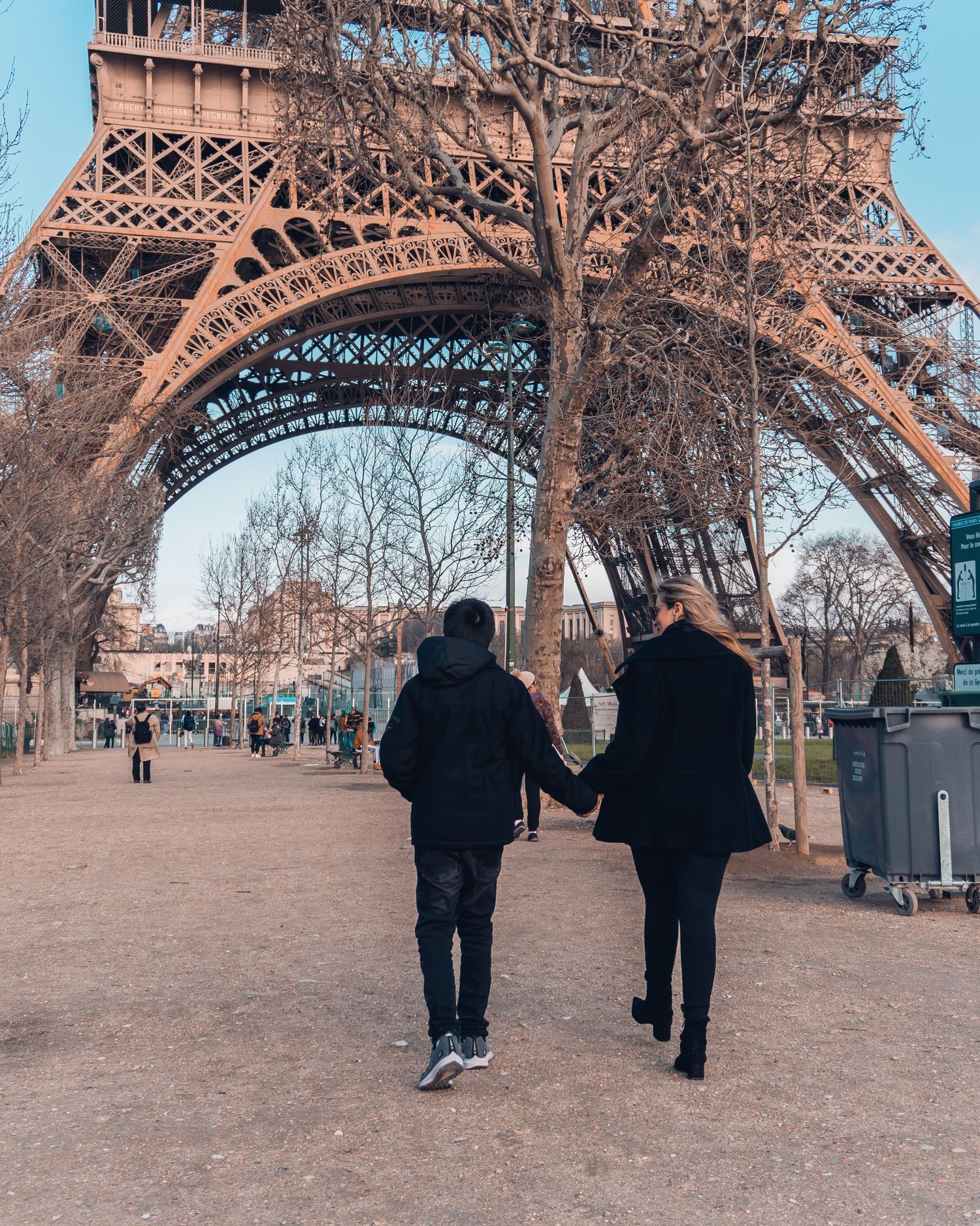 Silhouetted figures stand beneath the iconic latticed base of the Eiffel Tower in Paris on a cloudy day.