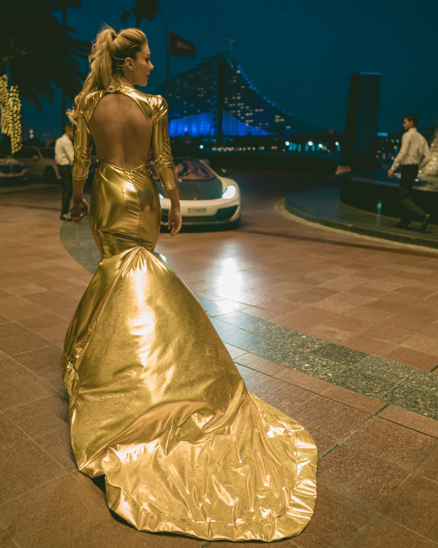A figure in a flowing gold evening gown poses dramatically against a dark nighttime cityscape background.