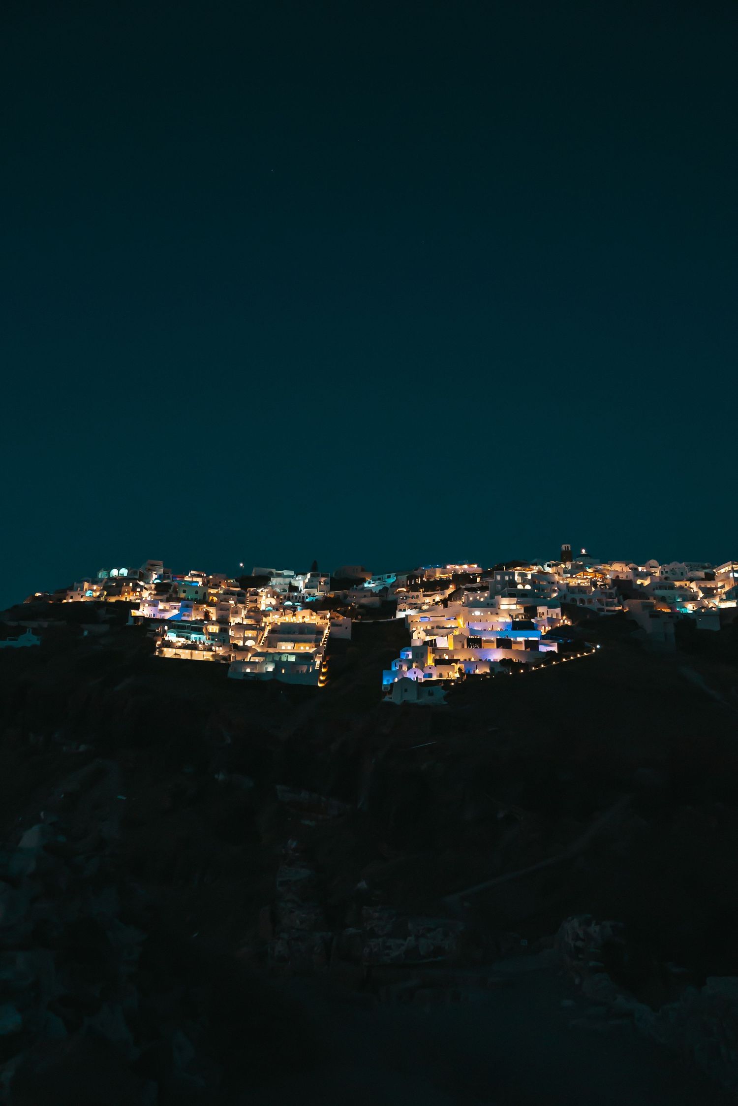 A hillside village illuminated at night with glowing lights scattered across the dark landscape against a deep blue sky.