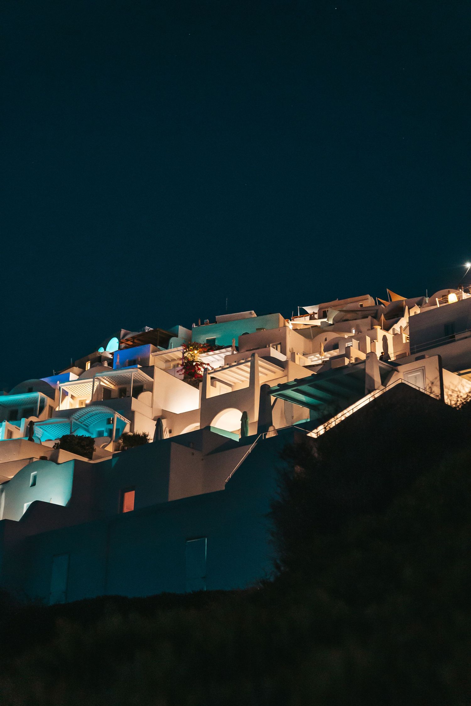 White-washed buildings with blue accents illuminate the hillside of Santorini at night creating a magical Mediterranean scene.