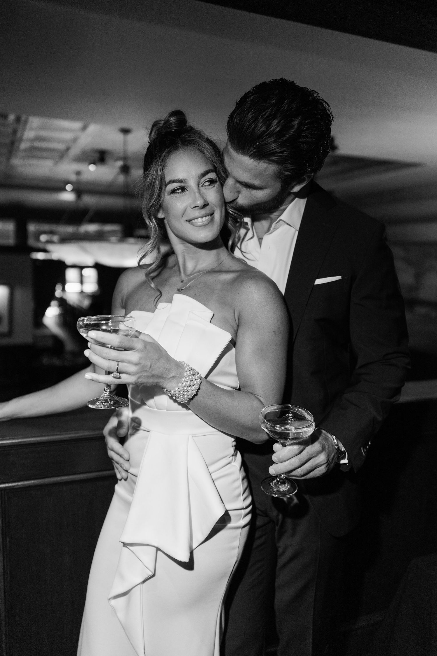 Elegant couple in black and white photo, woman in white strapless dress holding champagne, man in suit kissing her cheek.