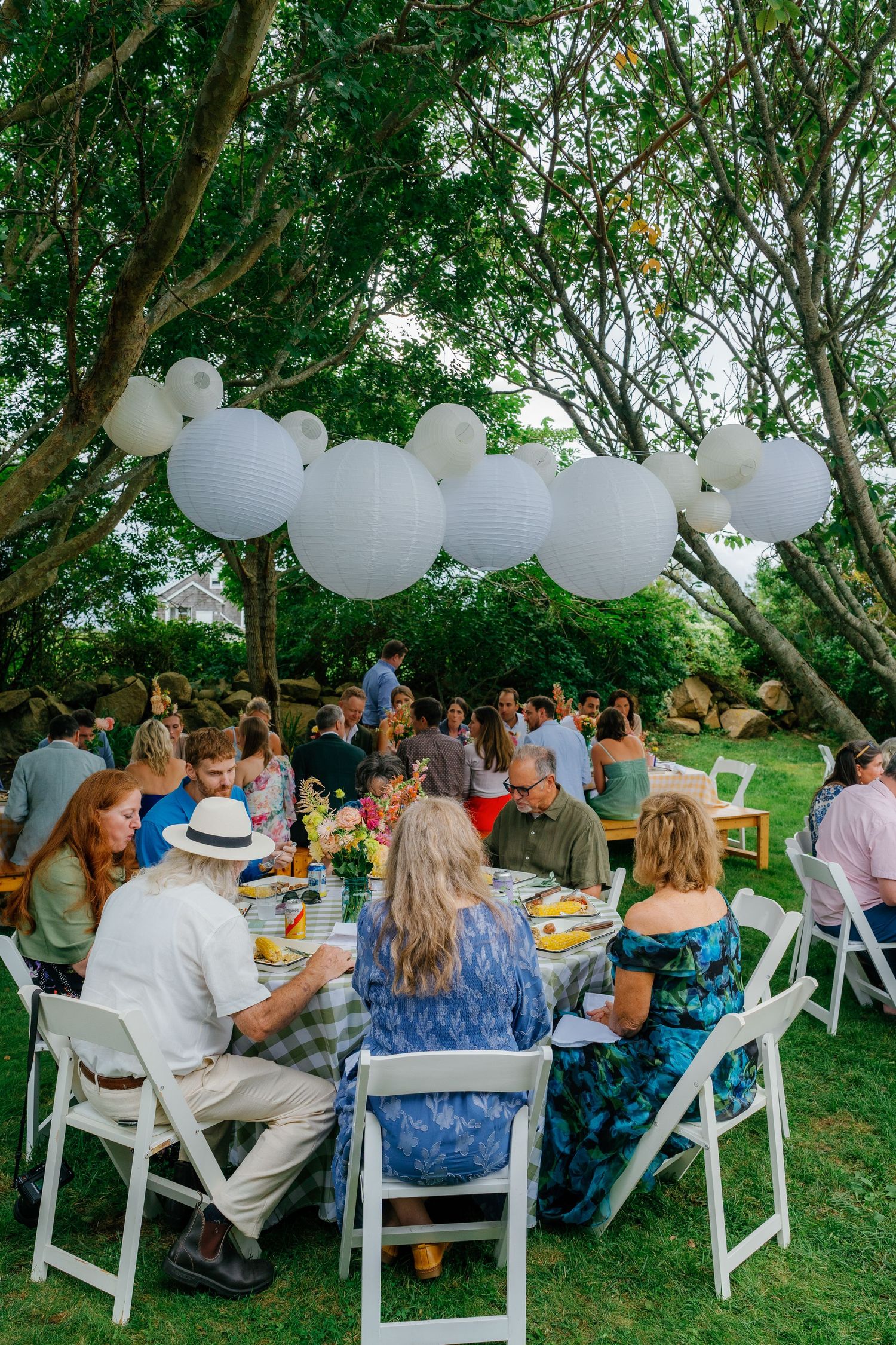 Outdoor garden party with guests dining at round tables under white paper lantern decorations hanging from trees.
