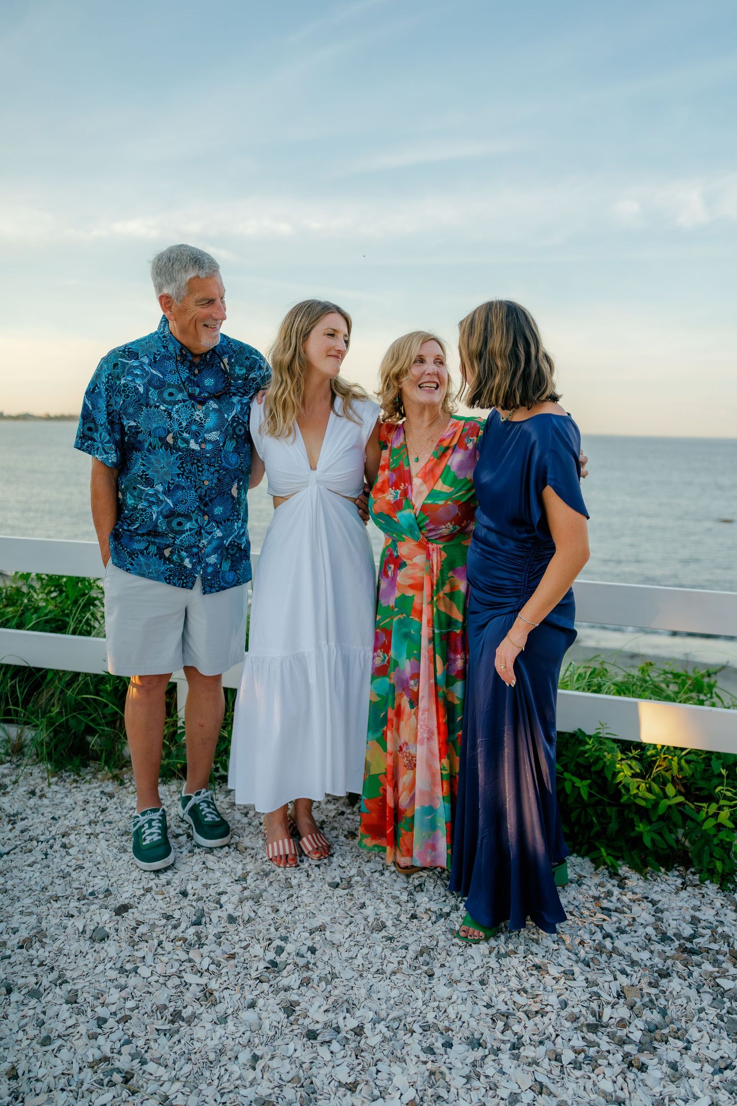 Four people in colorful outfits laughing together by a waterfront fence at sunset.