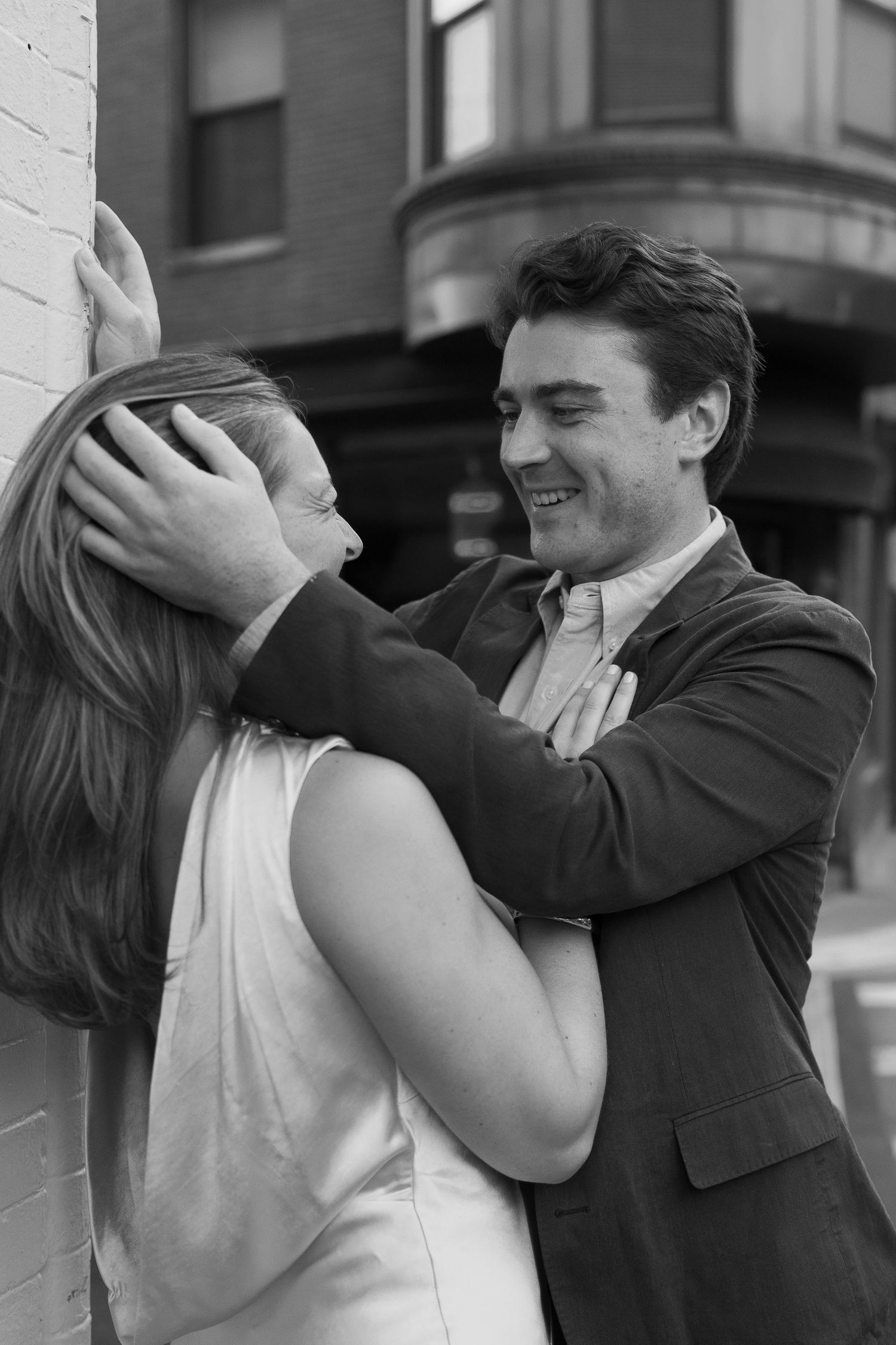 Black and white photo of a smiling man in a suit holding a woman against a brick wall in an urban setting.