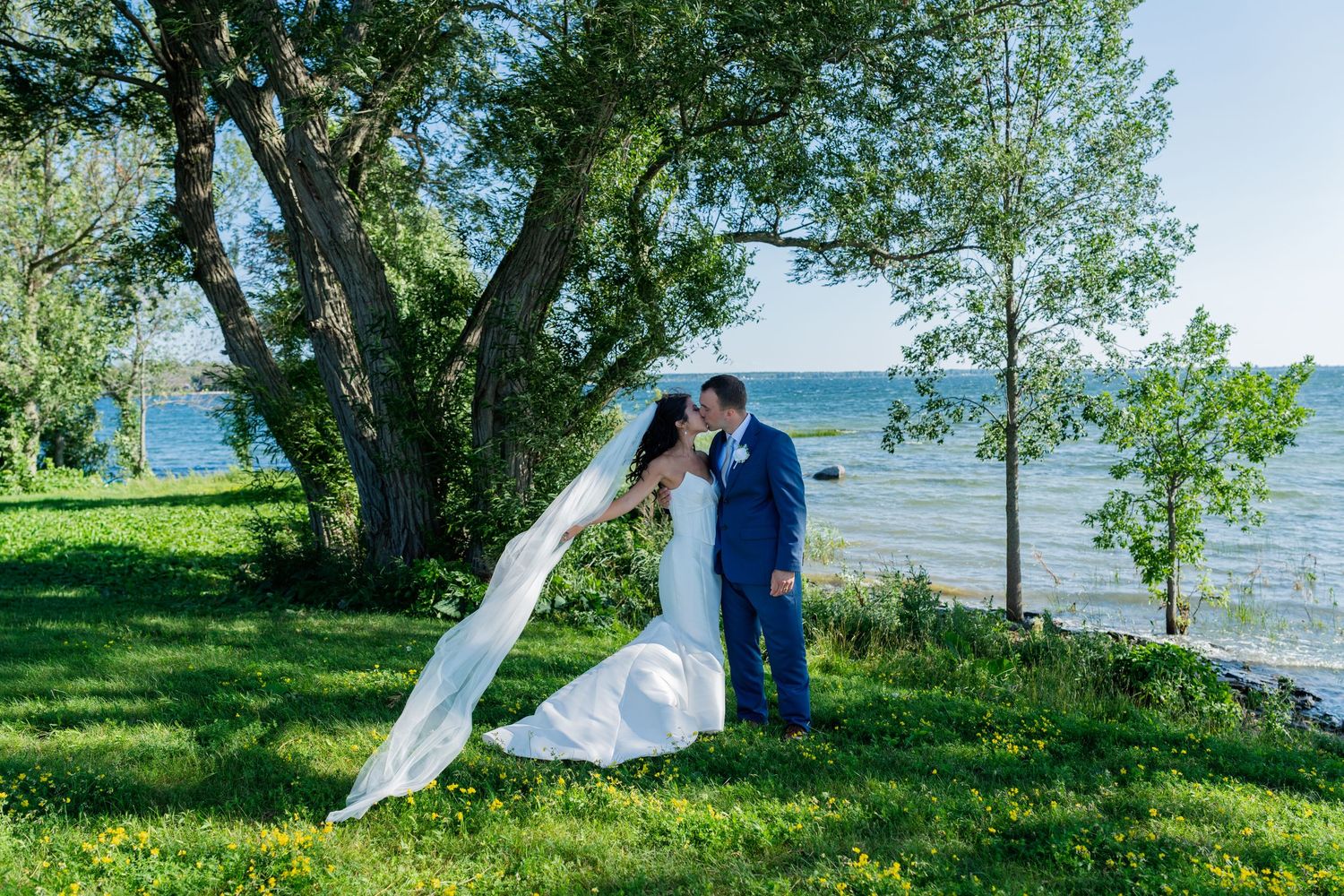 Bride and groom share a romantic kiss under a large tree by the waterfront on their wedding day.