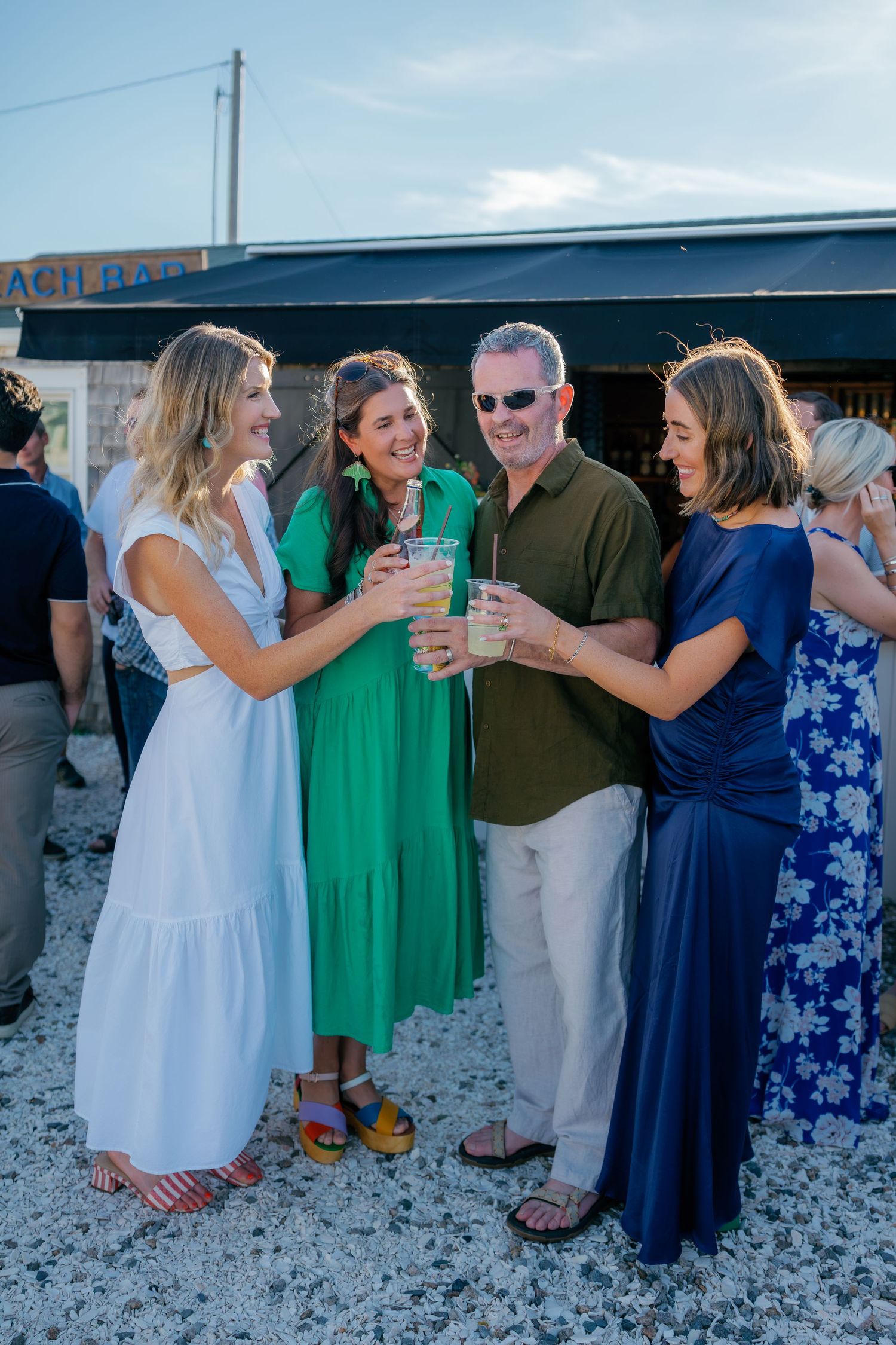 Four friends toast drinks and laugh together at an outdoor beach bar during a summer evening gathering.