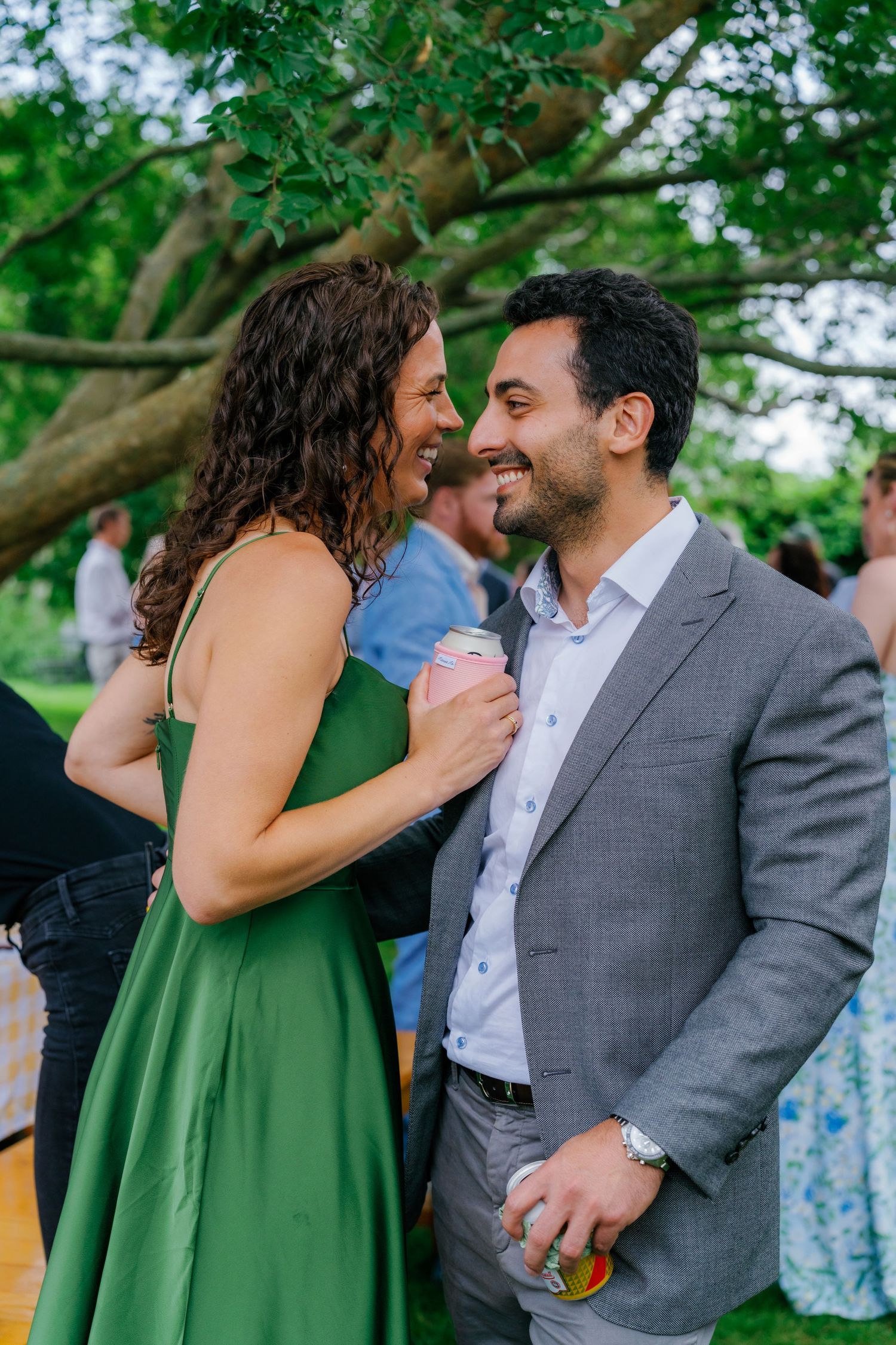 A couple in a green dress and gray suit smile at each other at an outdoor garden party event.