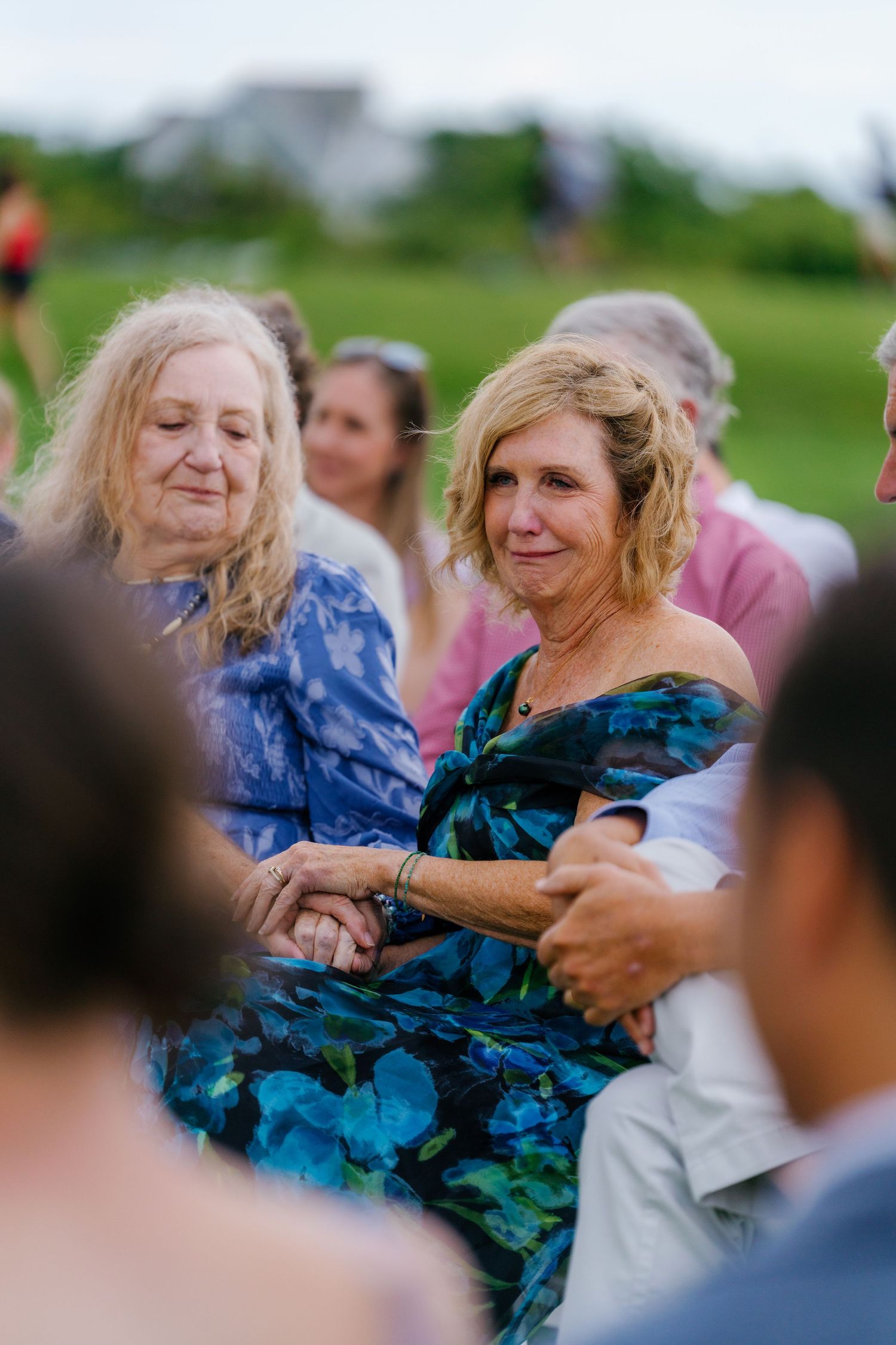 Two emotional women in floral dresses hold hands during an outdoor wedding ceremony on a lush green lawn.