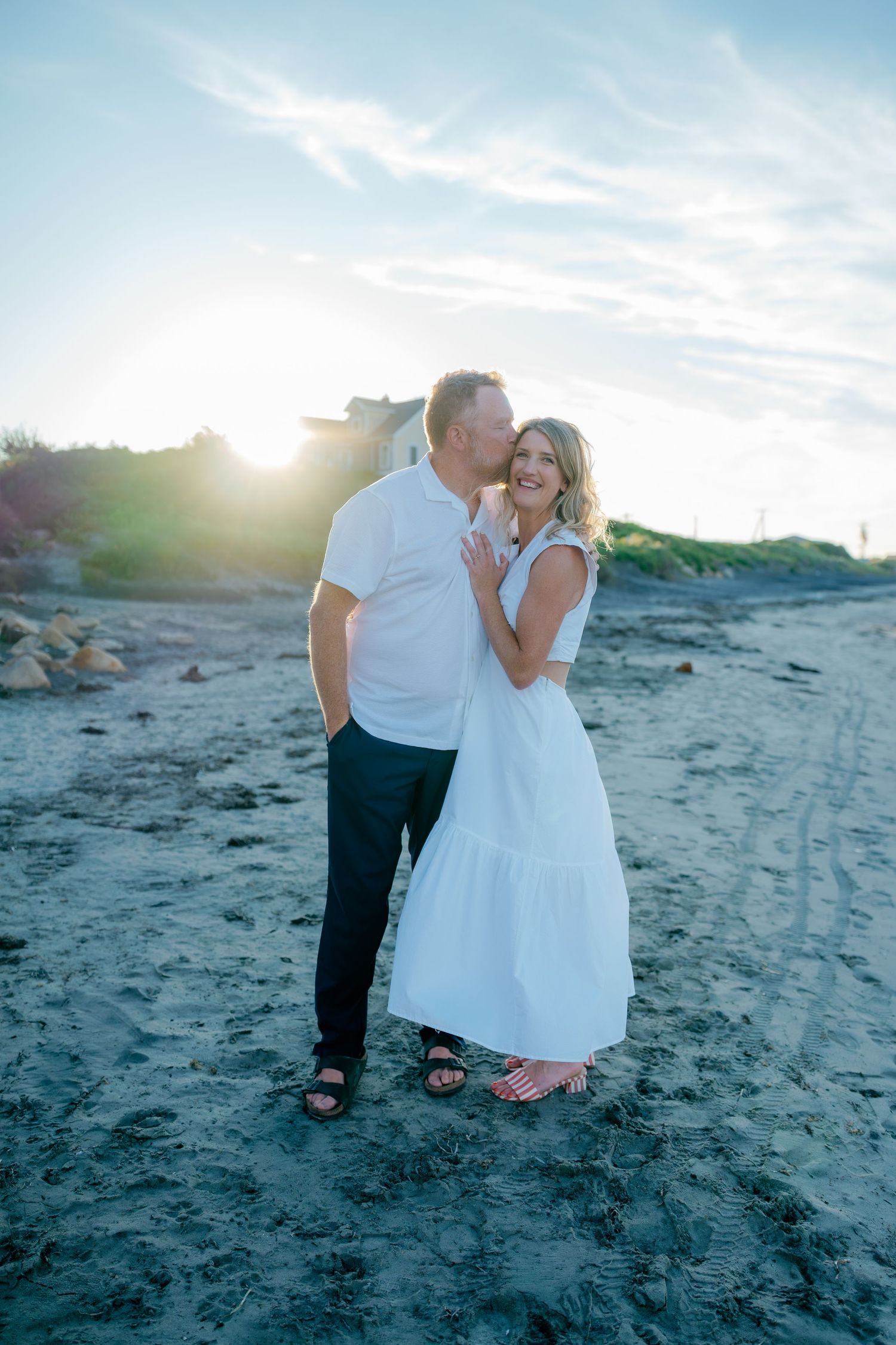 Couple embracing on a sandy beach at sunset, woman in white maxi dress and man in white shirt and black pants.