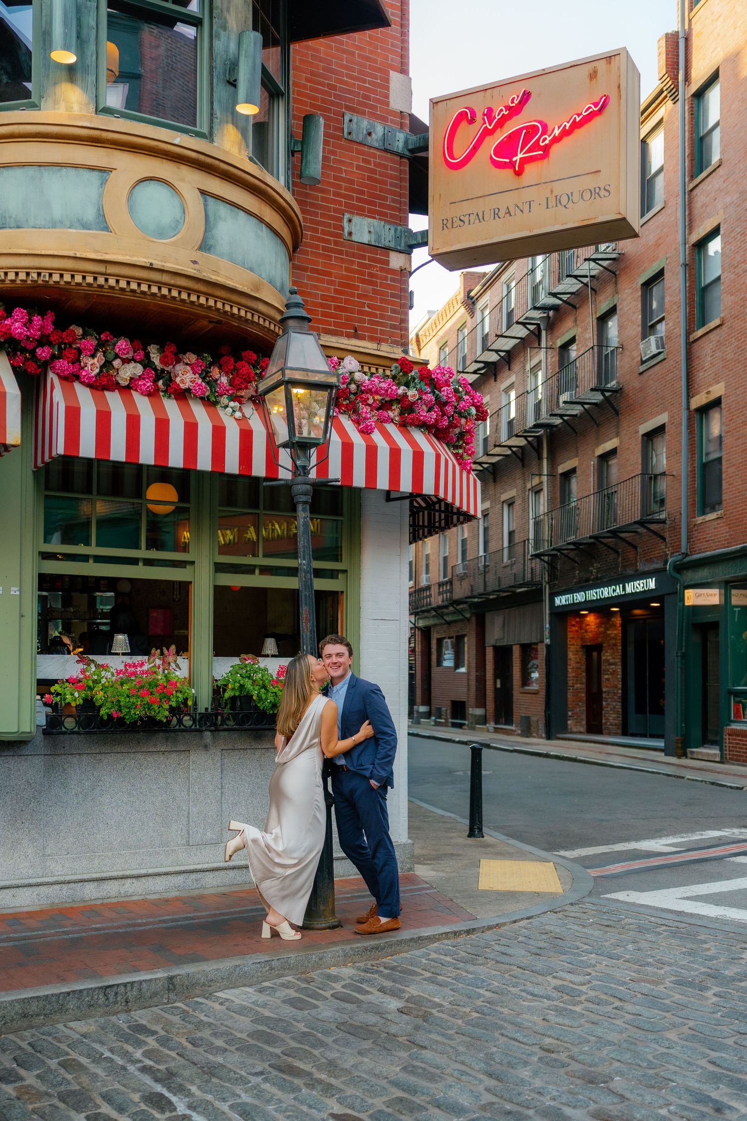 Couple embracing on cobblestone street corner near Chez Reno restaurant with red awnings and floral decorations.
