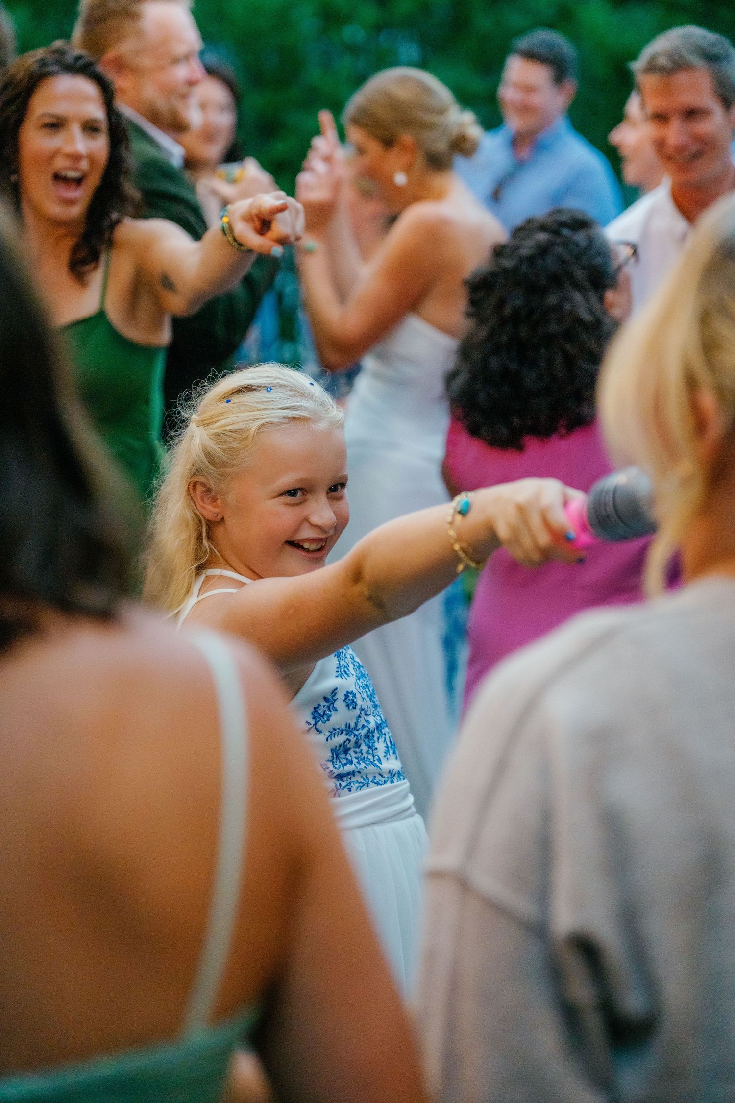 Young blonde girl smiling and pointing at a wedding reception surrounded by guests celebrating outdoors.