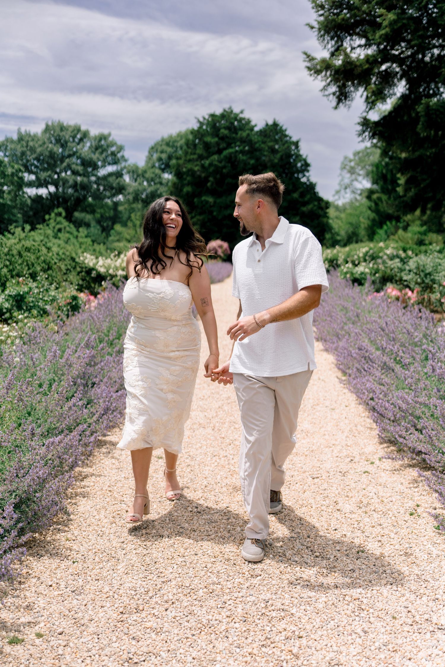 Couple holding hands and laughing while walking down a gravel path lined with lavender in a lush garden.