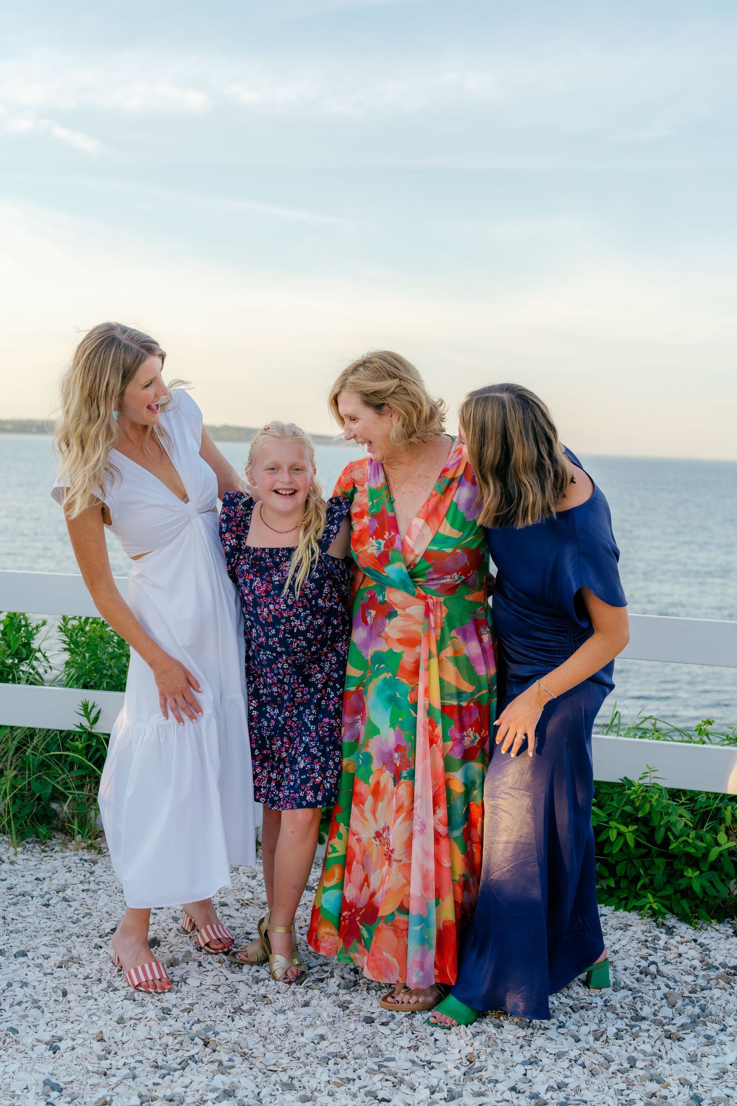 Four women and girls laughing together on a beachside deck at sunset, wearing colorful summer dresses.