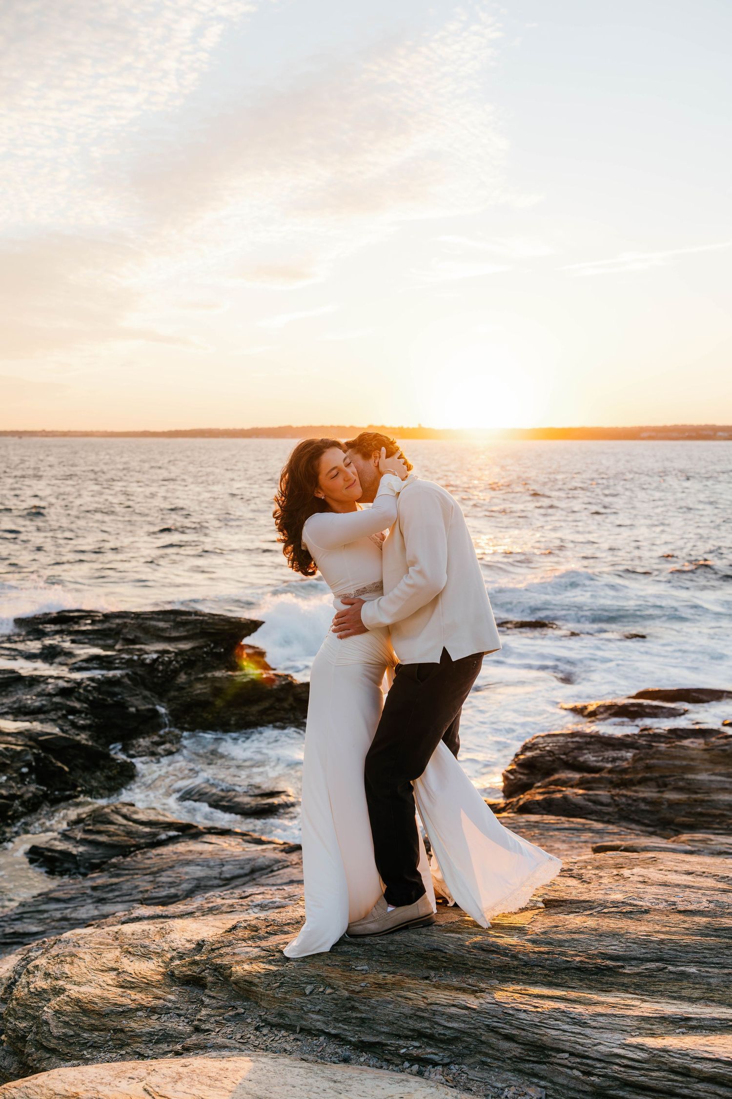 Bride and groom embrace on rocky shoreline at golden sunset, waves crashing behind them in romantic wedding portrait.