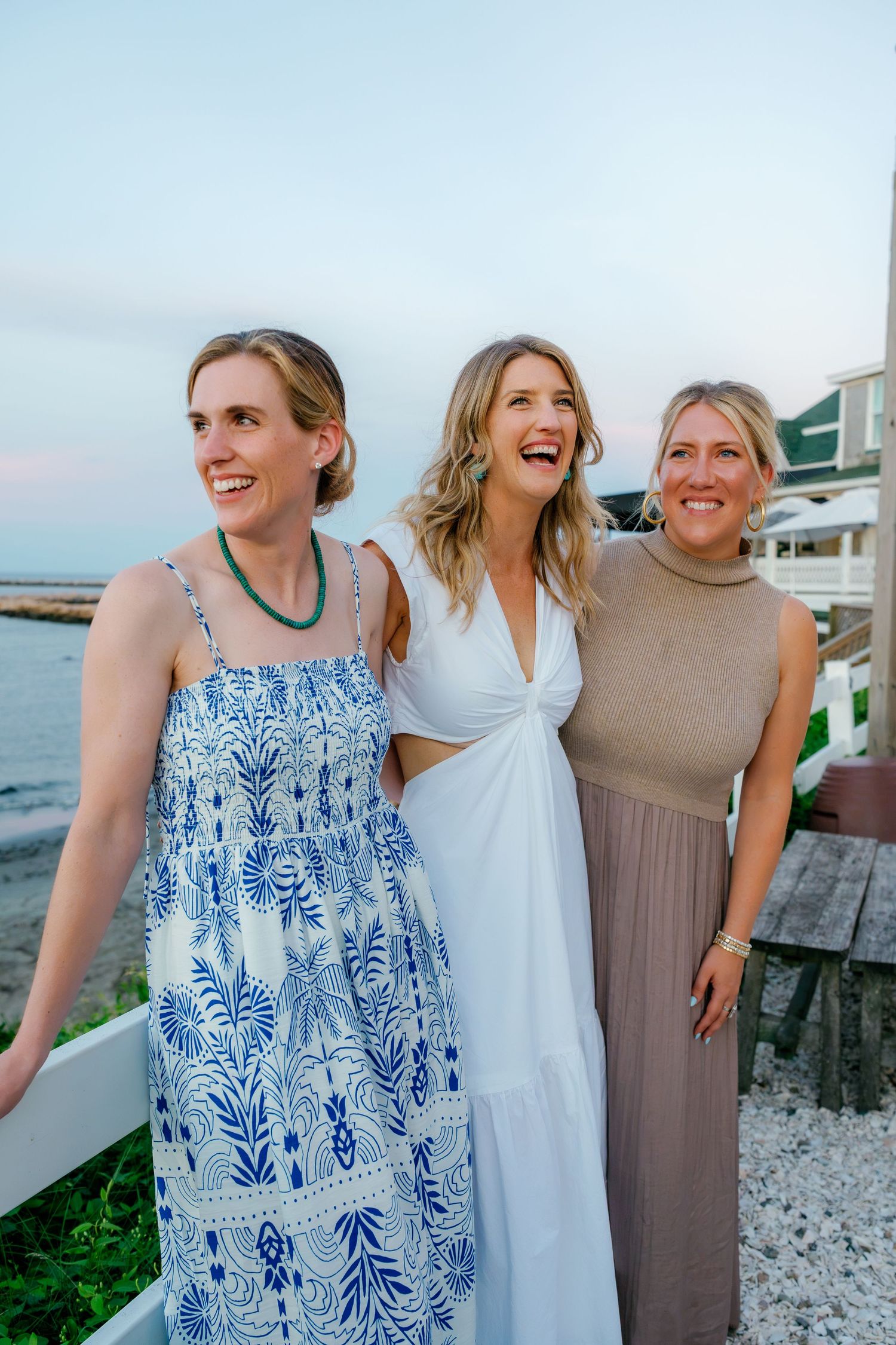 Three smiling women in summer dresses pose together on a coastal deck at sunset near the ocean.