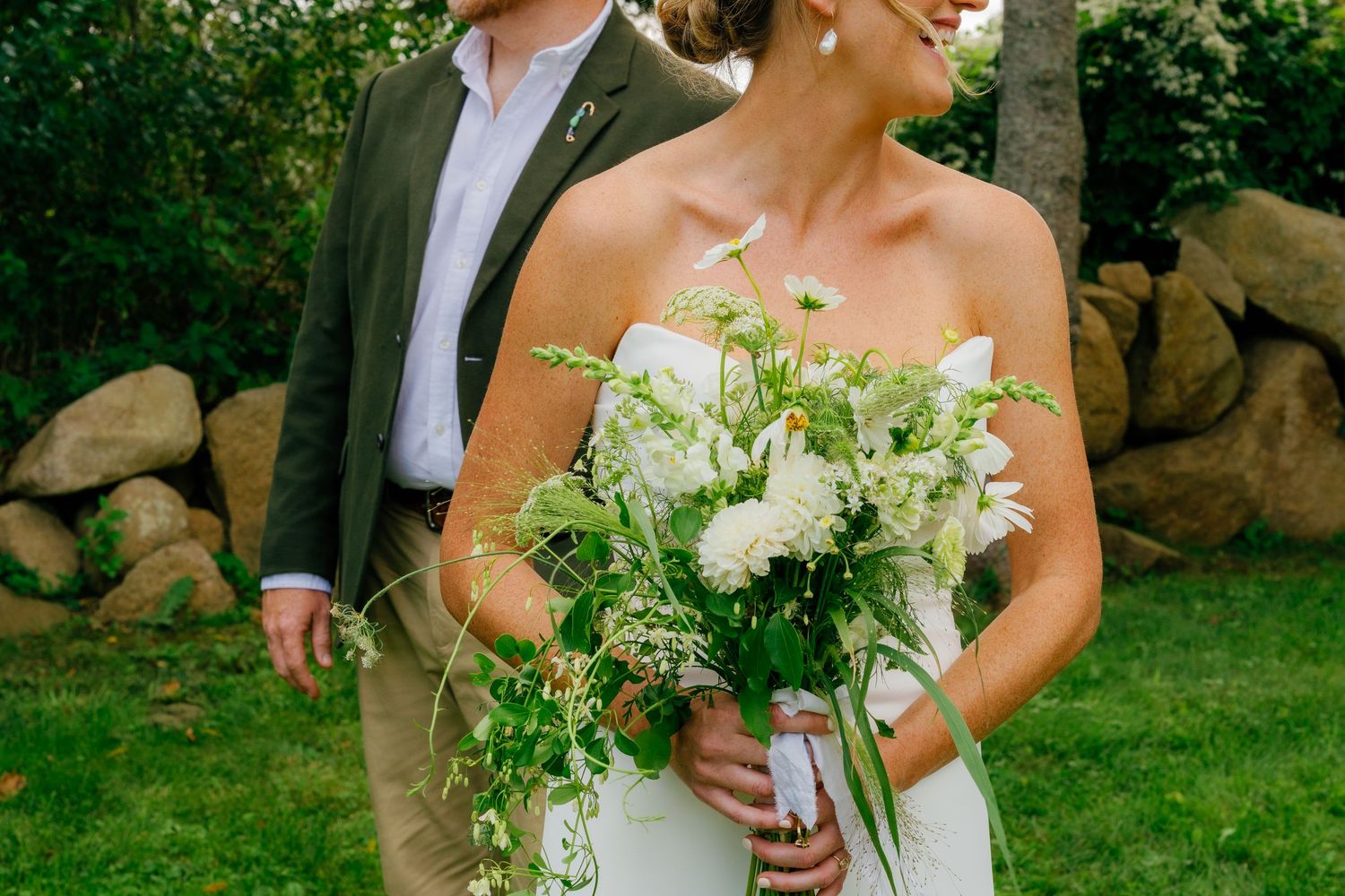 Bride holding lush white and green wedding bouquet stands beside groom in olive suit outdoors near stone wall.
