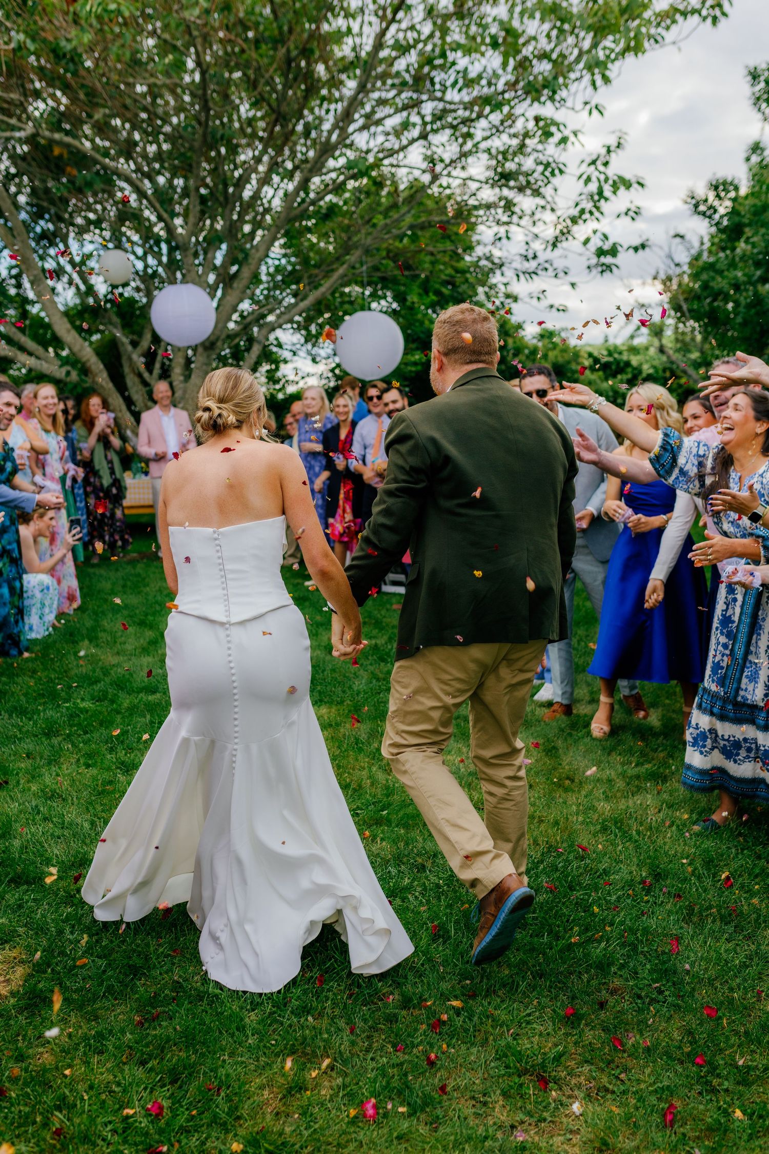 Bride and groom walk through cheering guests tossing confetti on green lawn at outdoor wedding celebration.