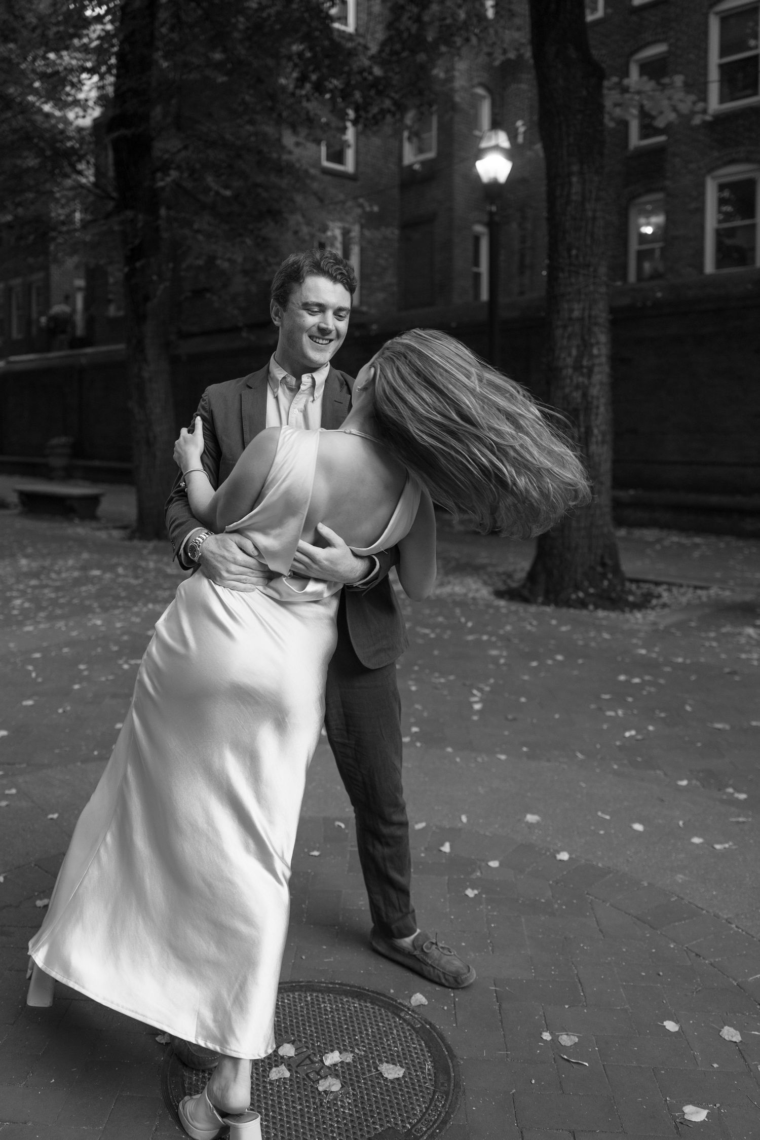 Black and white photo of a couple dancing outdoors at night, woman in elegant dress, man in suit, urban setting.