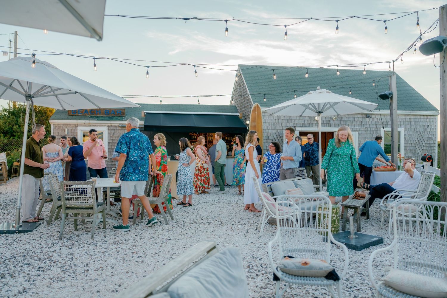 Outdoor beach party with guests mingling under string lights near white patio furniture and umbrellas at a coastal venue.