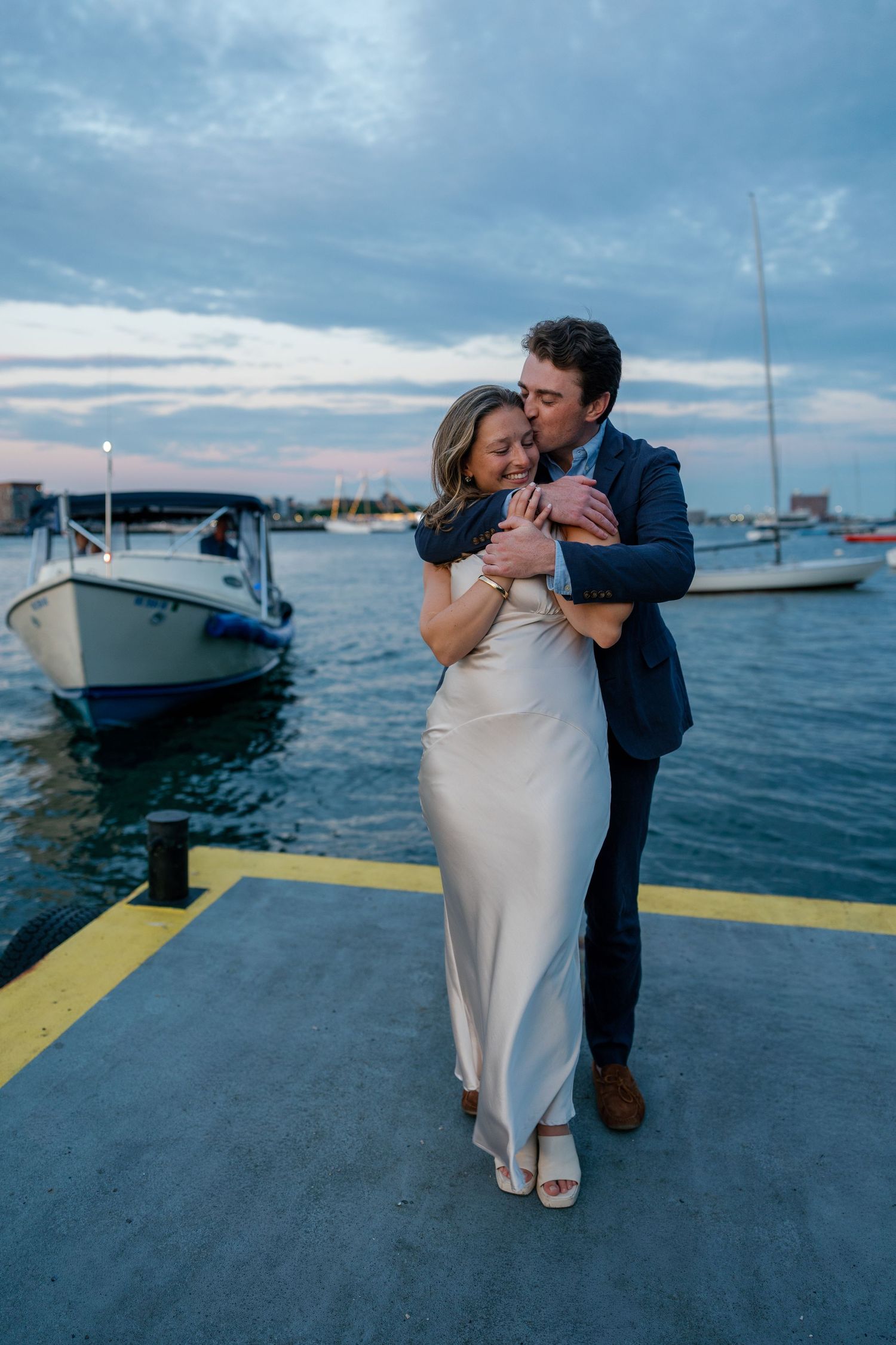 Couple embracing on a marina dock at dusk, woman in white satin gown, man in navy suit, boats in background.