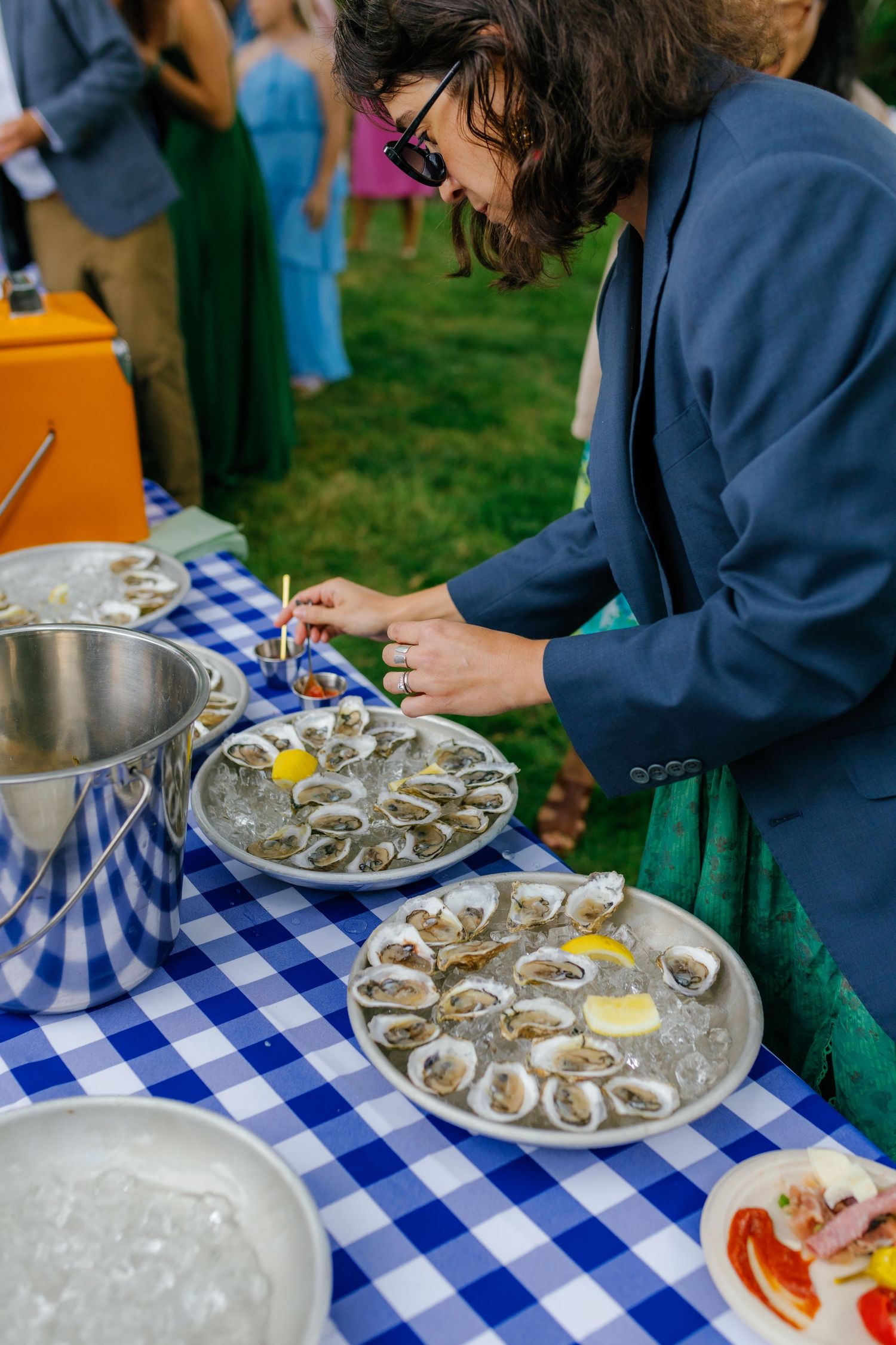 Person in blue blazer serving fresh oysters on ice at an outdoor event with blue checkered tablecloth.