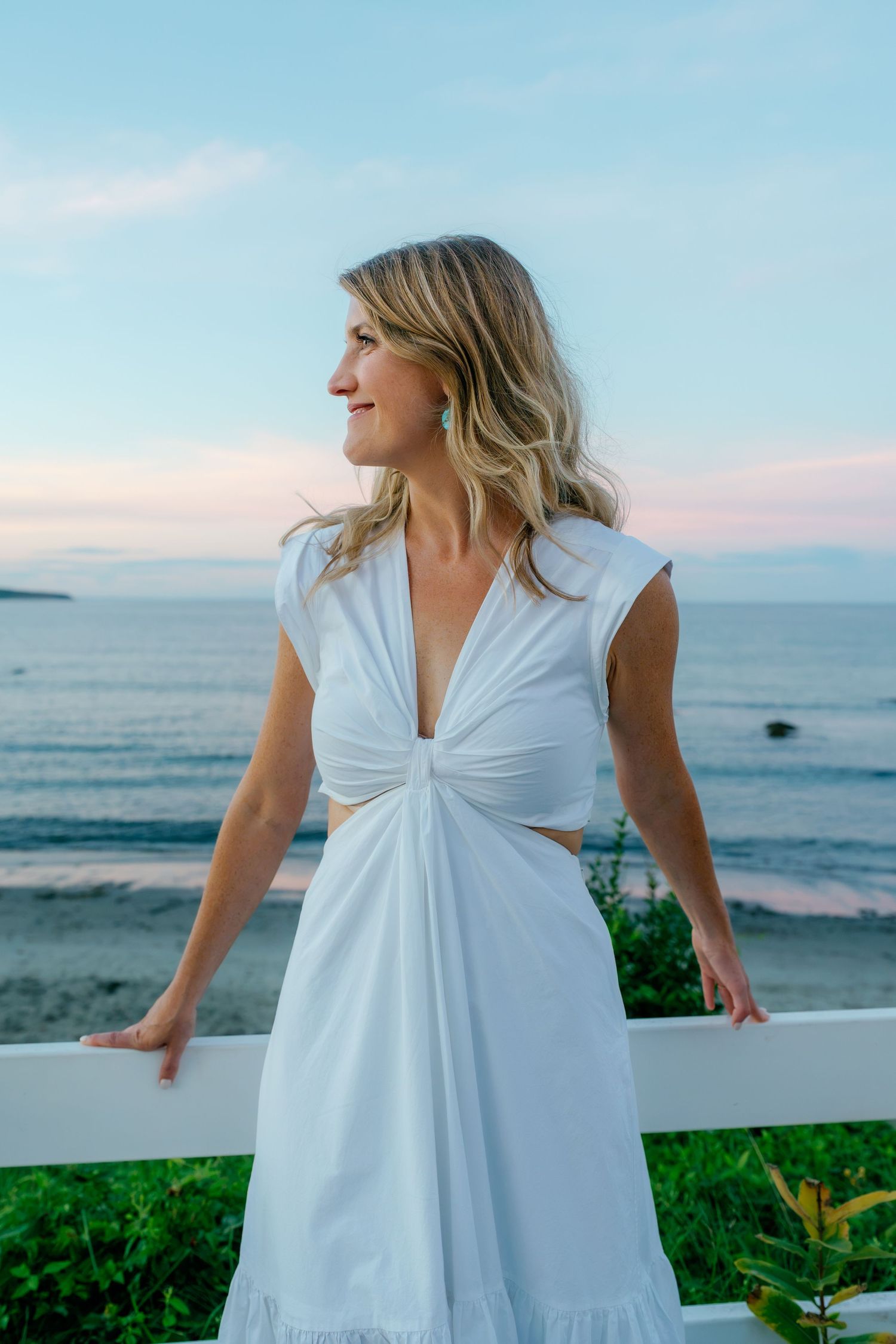 Blonde woman in white cut-out dress standing by a white fence with ocean view at dusk.