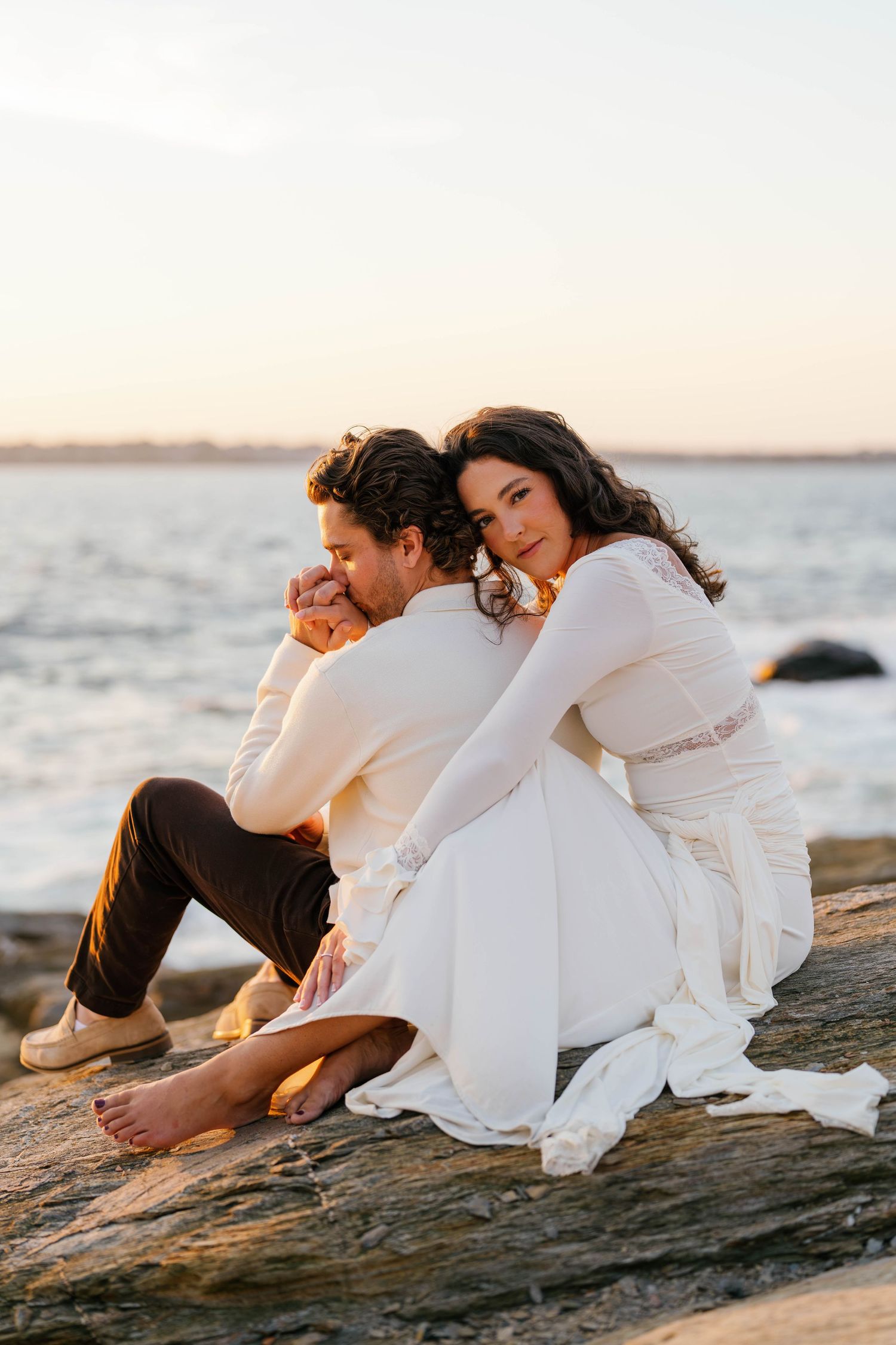 Couple in wedding attire sitting on rocks by the ocean at sunset, bride in white lace dress embracing groom from behind.