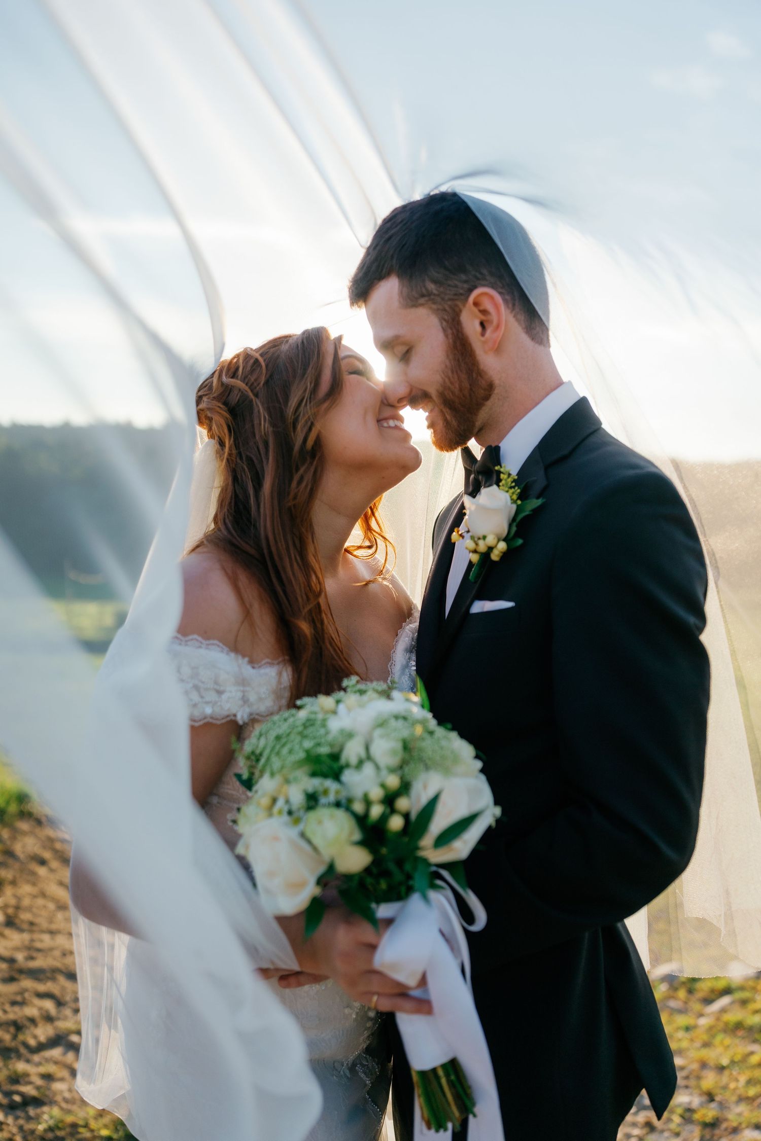 Bride and groom nearly kissing under flowing veil, holding white bouquet, groom wearing kippah, outdoor golden hour wedding photo.
