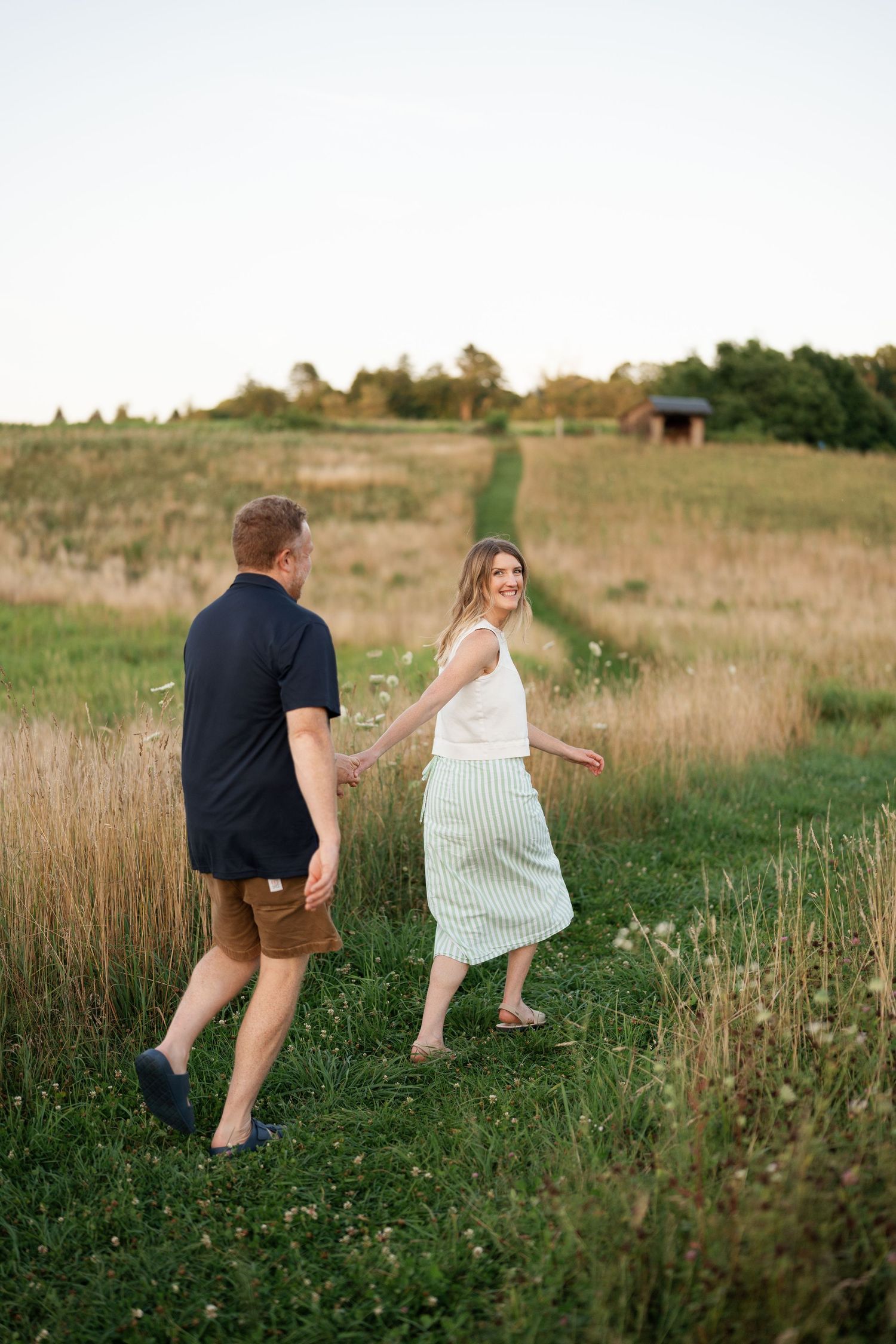 A couple walks hand-in-hand through a lush green meadow at sunset, the woman smiling back in a striped skirt.