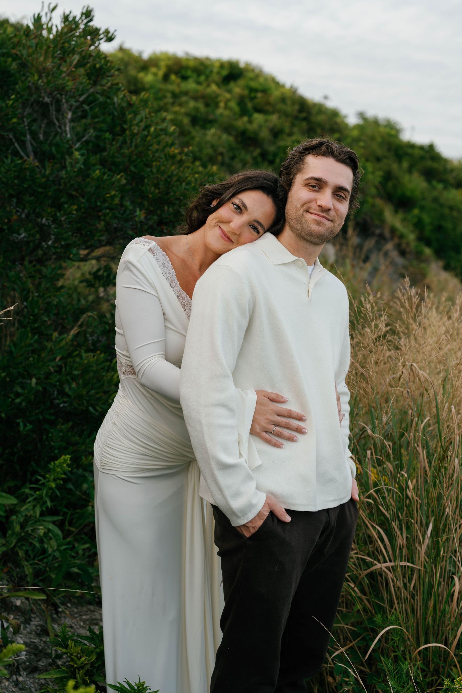 A couple in white outfits embraces outdoors, woman in gown leaning on mans shoulder amid lush greenery.
