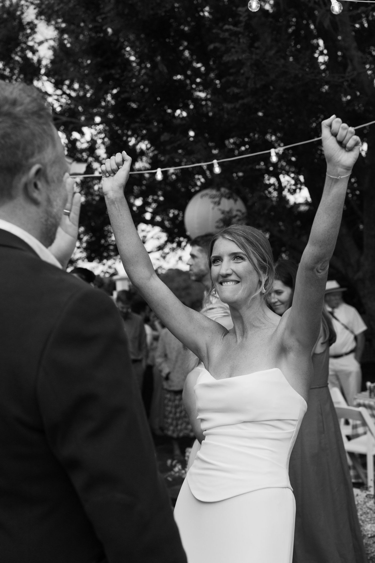 Black and white photo of a joyful bride in a strapless gown raising her arms in celebration at an outdoor wedding reception.
