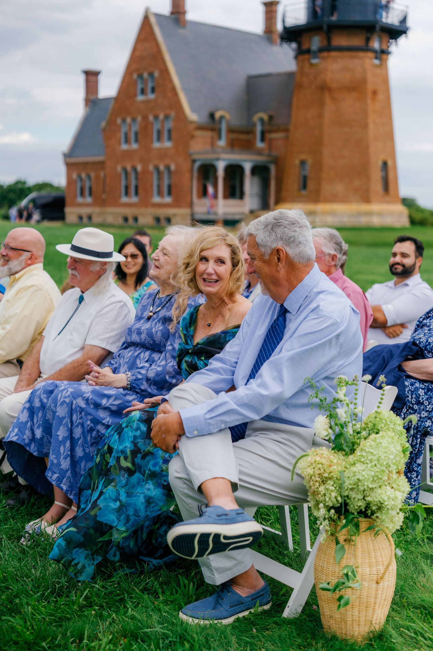 Guests seated outdoors at a wedding ceremony on a green lawn with a historic brick lighthouse building in the background.