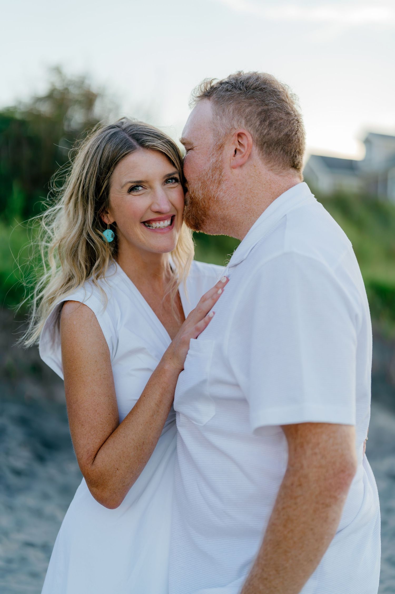 Couple in white outfits sharing a tender moment on a sandy beach at sunset, man kissing woman's cheek.